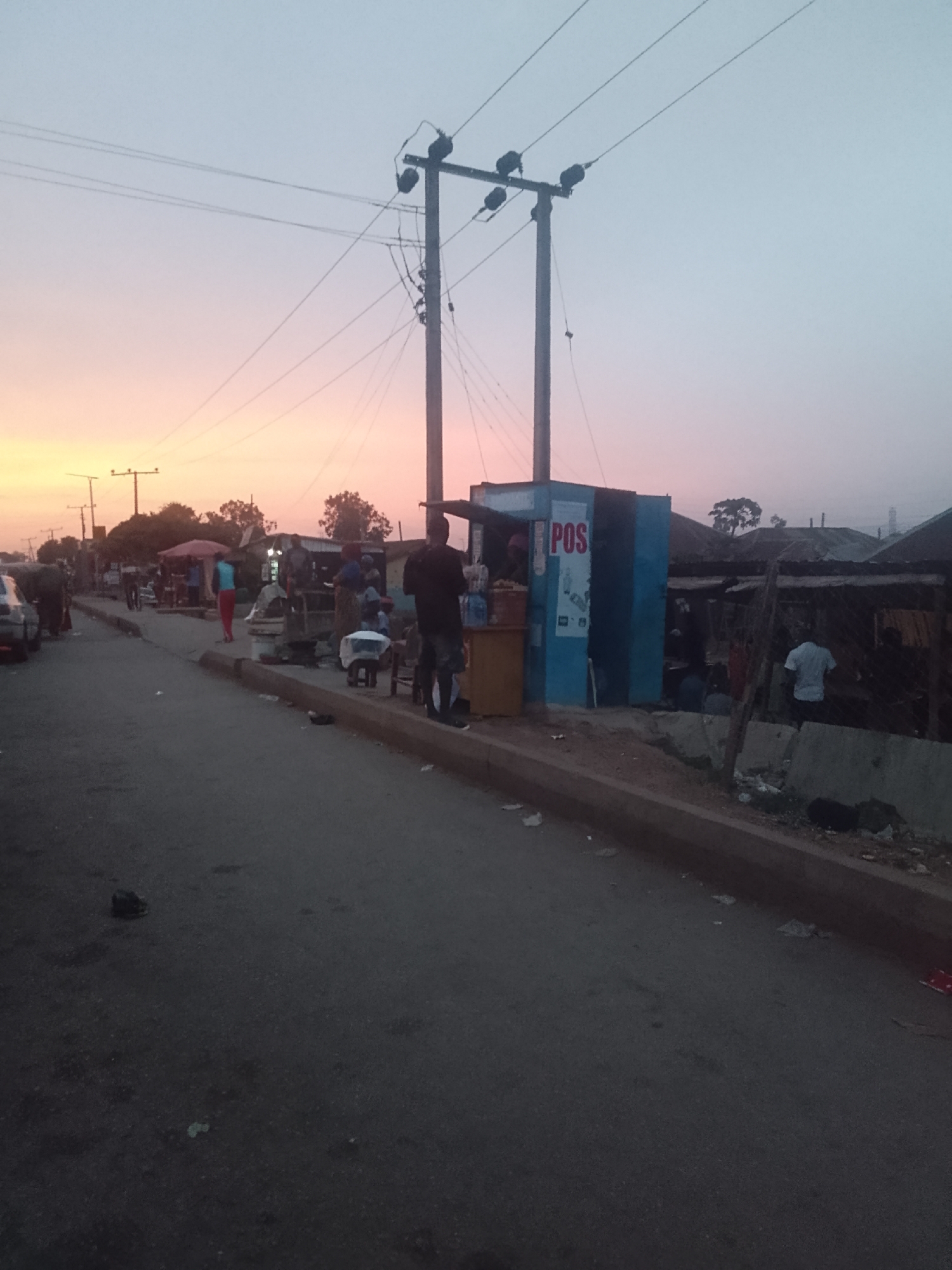 The image depicts a street scene in Bukuru, Nigeria at dusk. The sky exhibits a gradient of orange and pink hues as the sun sets. A two-lane road occupies the foreground, with a raised concrete curb separating it from the sidewalk. On the sidewalk, several people are gathered, and there are small structures that appear to be small businesses or kiosks. One blue structure has "POS" printed on its exterior. Power lines and utility poles are visible, with one pole located next to the "POS" kiosk. Litter is scattered on the ground near the structures.