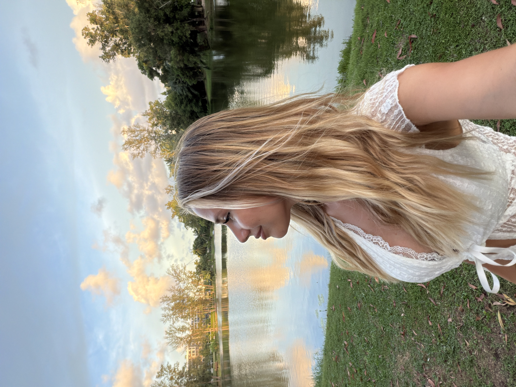 A young woman with long, blonde, wind-swept hair is captured in a close-up, side profile, looking down towards a body of water. She is wearing a white, lace-trimmed top. The setting appears to be outdoors, with a grassy area in the foreground and a calm lake or pond behind her. The water reflects the sky and surrounding trees, suggesting a natural environment. In the background, there are lush green trees and some distant buildings or structures. The sky is a mix of blue with soft, warm-toned clouds, indicating it might be late afternoon or early evening, possibly during sunset, given the color of the clouds. The overall mood seems serene and peaceful. There is no visible text in the image.