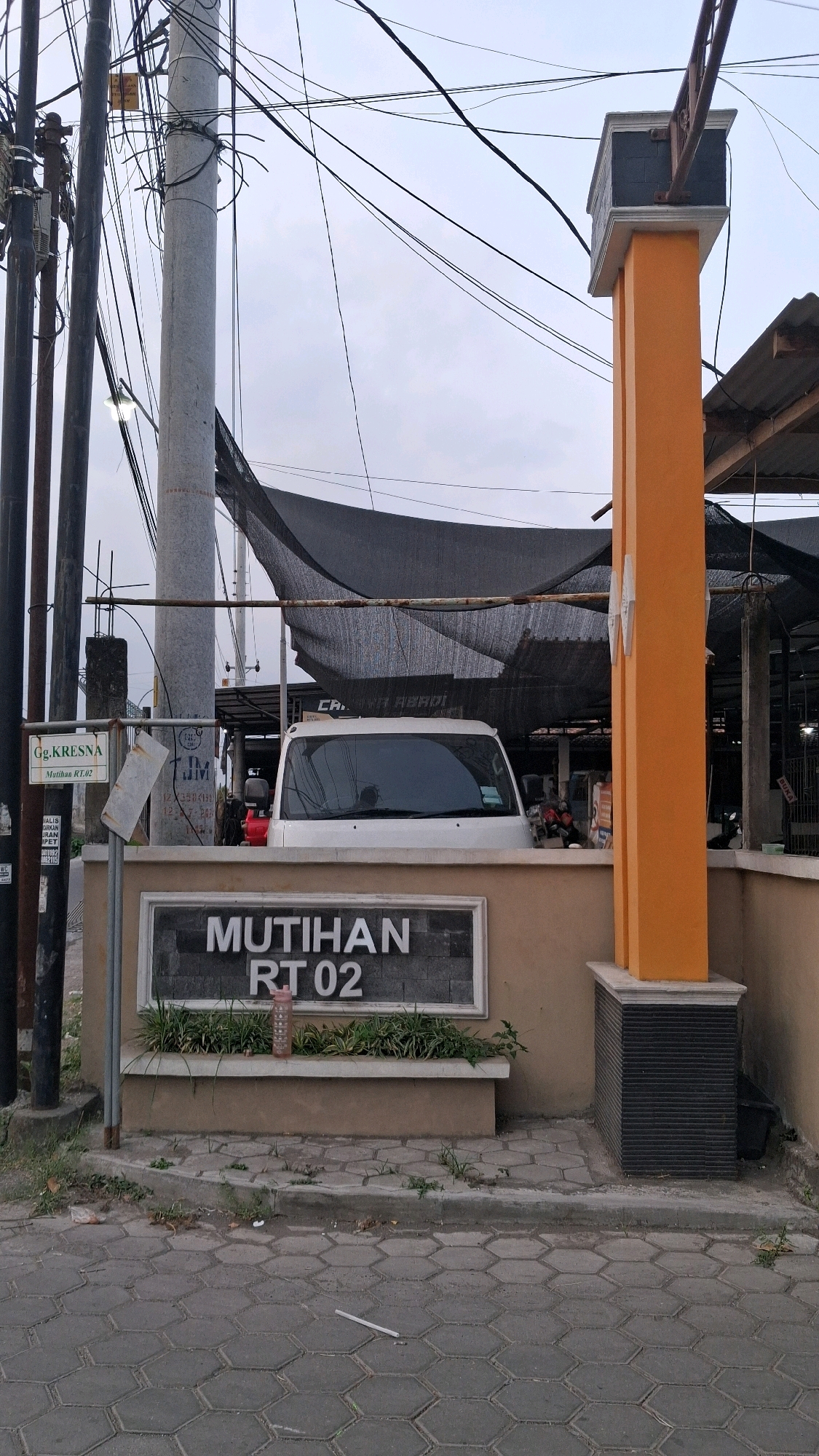 A white van is parked behind a sign that reads "MUTIHAN RT 02". The sign is on a wall in front of an orange pillar. There are power lines overhead and a black tarp is stretched above the van. The street is paved with hexagonal bricks.