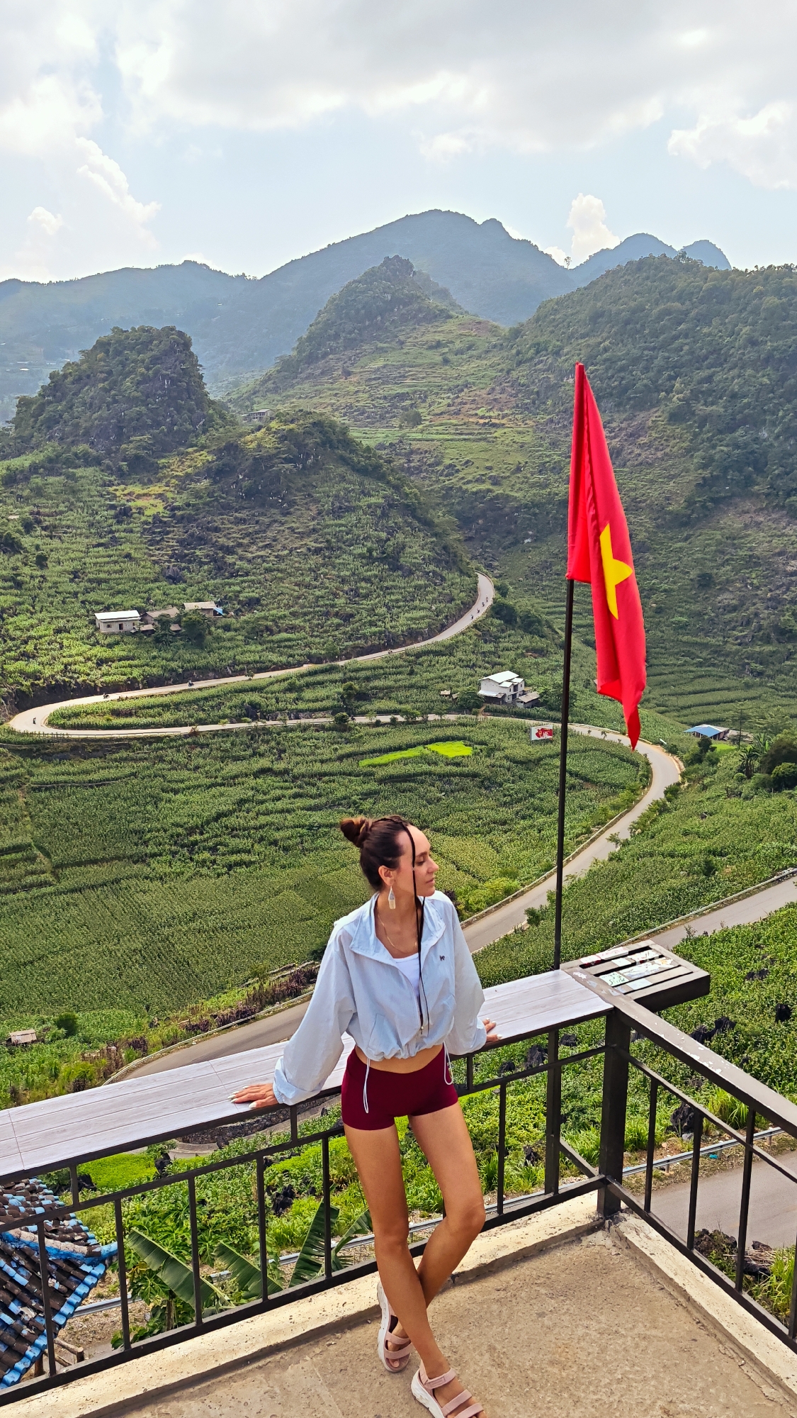 A woman is standing on a balcony overlooking a mountainous landscape in Lũng Phìn Commune, Vietnam. She is wearing a light blue jacket, red shorts, and pink sandals. Her hair is tied up in a bun. She is leaning on the balcony railing, looking off into the distance. A Vietnamese flag is positioned on a pole nearby. The background features rolling hills covered in green vegetation, a winding road, and scattered houses. The sky is partly cloudy.