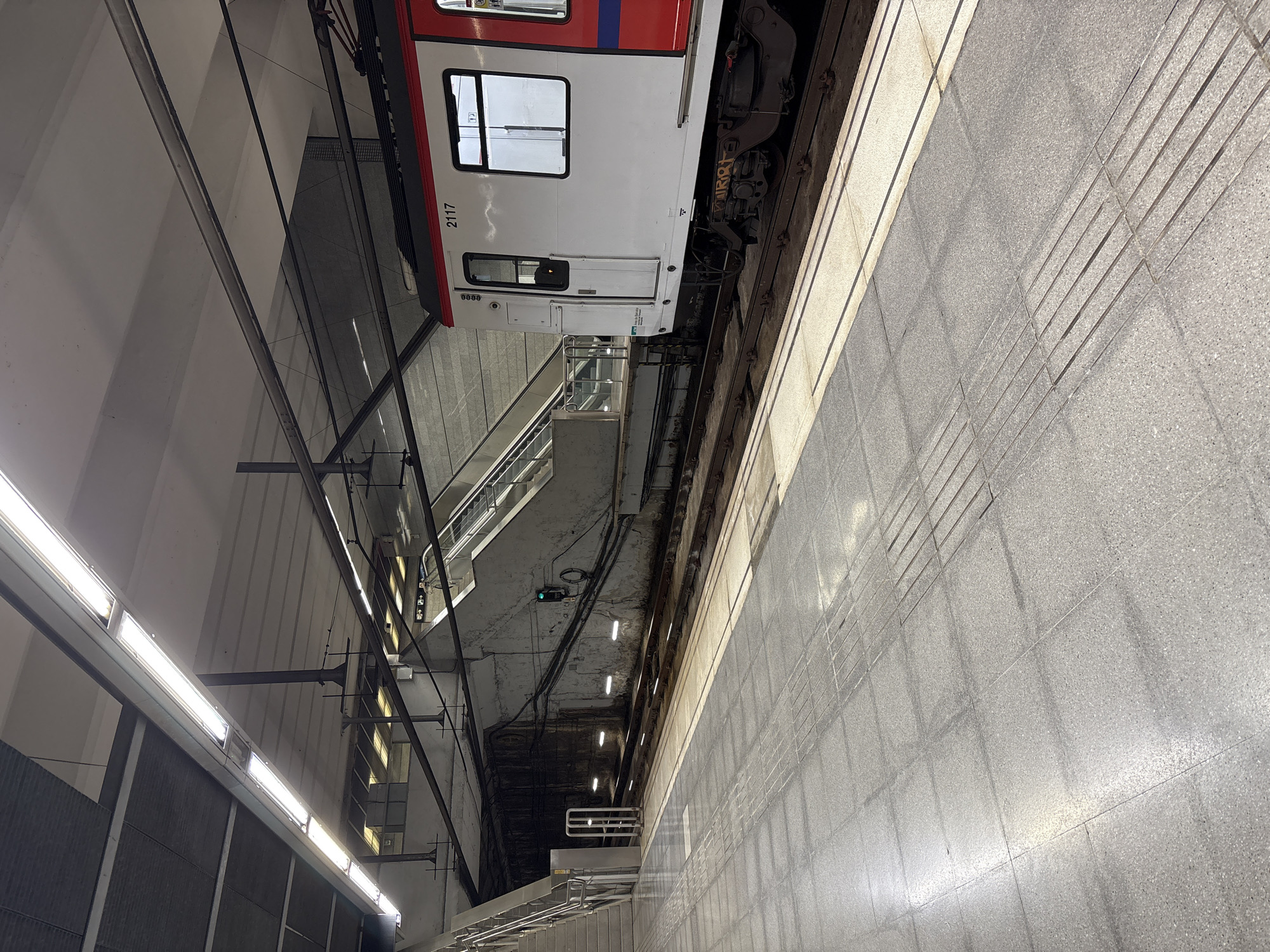The image depicts a subway station platform with a train stopped at the platform. The platform is made of grey tiles with tactile paving for the visually impaired. To the left, there are stairs leading up. In the background, there is a tunnel entrance and escalators. A white and red subway train, numbered 2117, is parked alongside the platform. The station is well-lit by fluorescent lights. The environment appears clean and well-maintained. There are no people visible in the image.