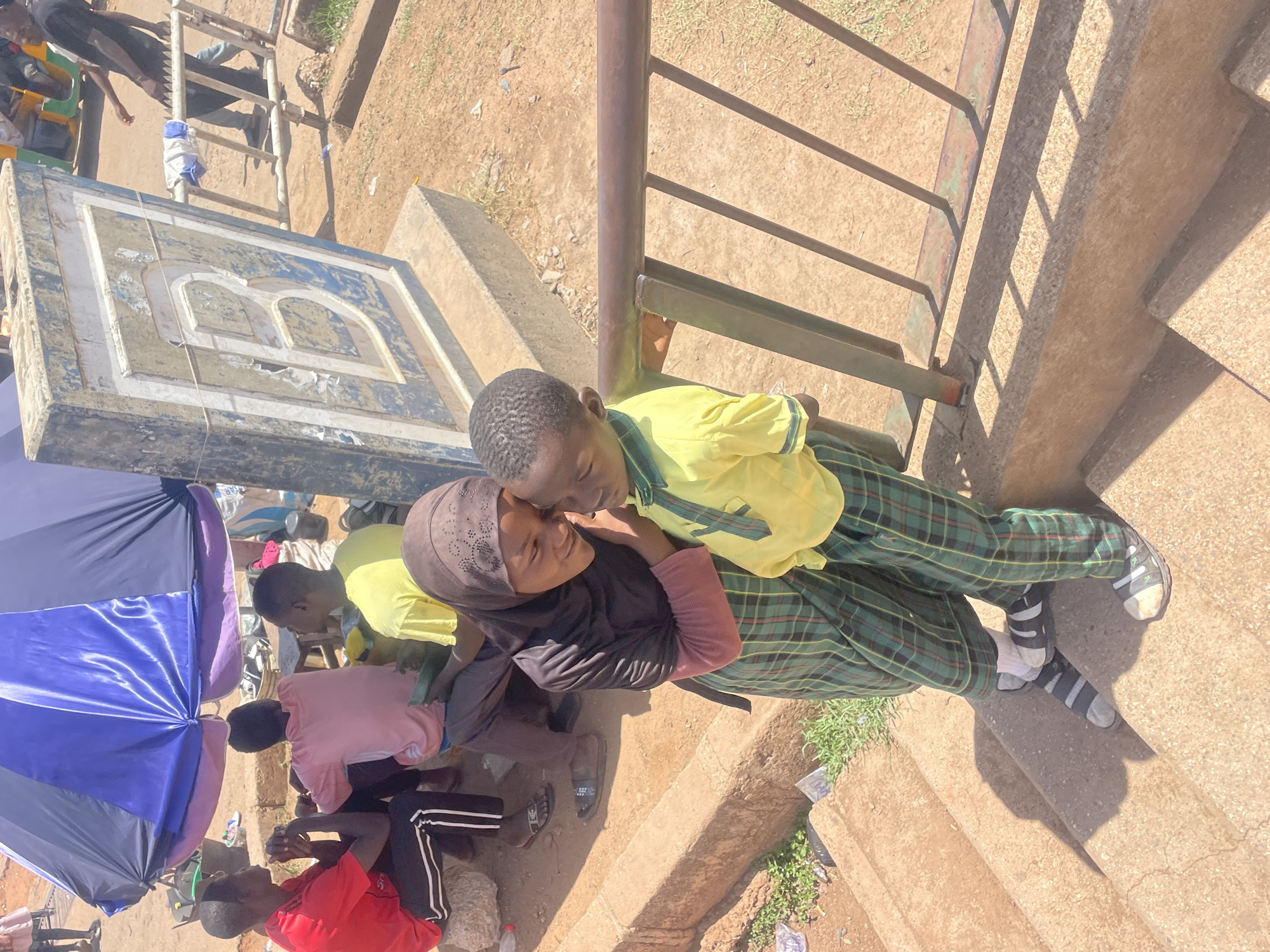 Two school-aged children, one in a hijab, are nuzzling noses on stairs with a metal railing, while others are seated nearby under an umbrella. The children are wearing school uniforms. The scene is situated outdoors. A signpost with a large letter "B" is visible in the background. The location is Gura Top, Nigeria.