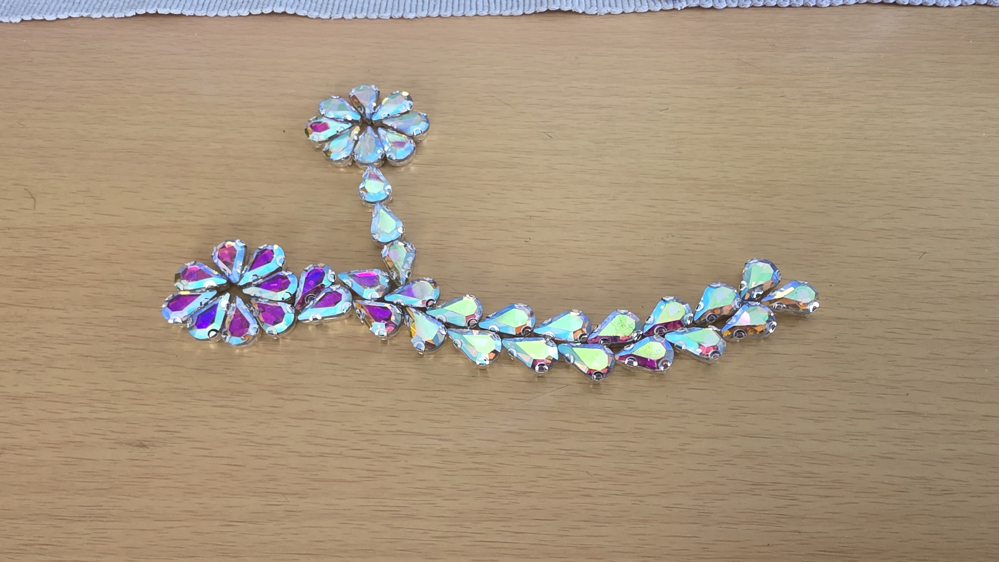 The image shows an arrangement of iridescent crystal embellishments on a wood-grain surface. The embellishments are mostly tear-drop shaped, arranged in two floral formations connected by a curving stem-like structure also composed of tear-drop crystals. The floral formation at the top of the image is lighter in color while the one at the bottom left is darker, with hints of pink and purple. The "stem" curves to the right side of the image. The background is a light brown wood surface with minimal texture. A portion of a white cloth with a subtle patterned edge is visible at the very top of the frame.