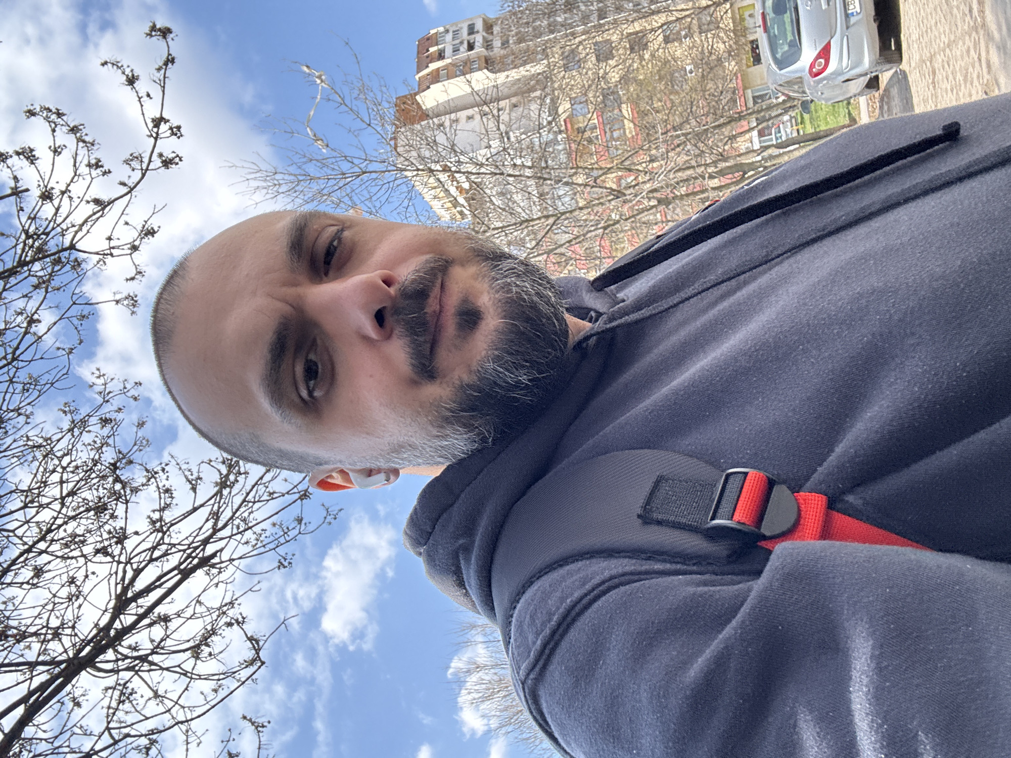 A male subject is depicted in a close-up, low-angle selfie taken outdoors under a bright blue sky with scattered white clouds. The man has a bald or closely shaved head, a dark full beard with graying at the temples, and is wearing a dark hooded sweatshirt. A white earbud is visible in his right ear. A dark gray backpack strap with a red webbing and black buckle is visible over his right shoulder. In the background, bare tree branches are present. To the right, a multi-story apartment building with a light-colored facade and red-orange detailing is visible, partially obscured by additional bare trees. A silver Opel Corsa hatchback is parked on the right, facing away, displaying a partial license plate that includes "CB," indicating Sofia, Bulgaria.