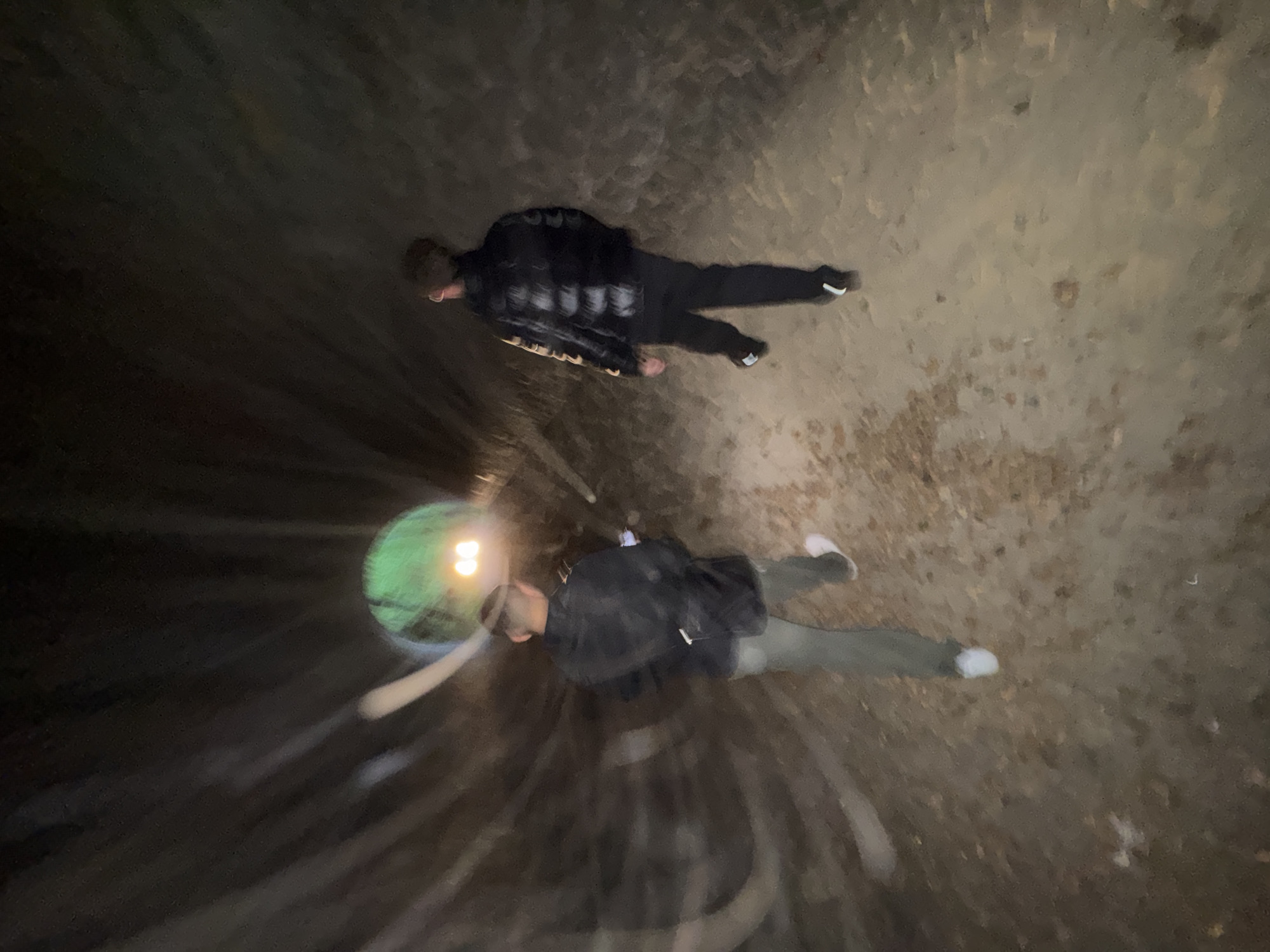 The image provides an overhead view of two individuals exploring a dark, enclosed tunnel or large pipe in Bures-sur-Yvette, France. The primary light source is a bright, green-yellow headlamp worn by the person in the foreground, creating significant lens flare and a radial blur effect that suggests movement or a long exposure.
The individual further away is dressed in a plaid shirt over a dark garment, dark pants, and white-soled shoes. The person closer to the camera wears a dark jacket or sweatshirt, light-colored pants, and white shoes, and appears to be carrying a dark bag or backpack. Both figures are oriented as if moving forward through the space.
The environment is dim, with dark, seemingly damp, curved walls and a textured, earthy or concrete floor. The complete reliance on artificial light underscores the subterranean or deeply enclosed nature of the location. No specific text is visible within the image, and the subjects' expressions are not discernible. The overall impression is of an exploratory activity within a confined and dimly lit passage.