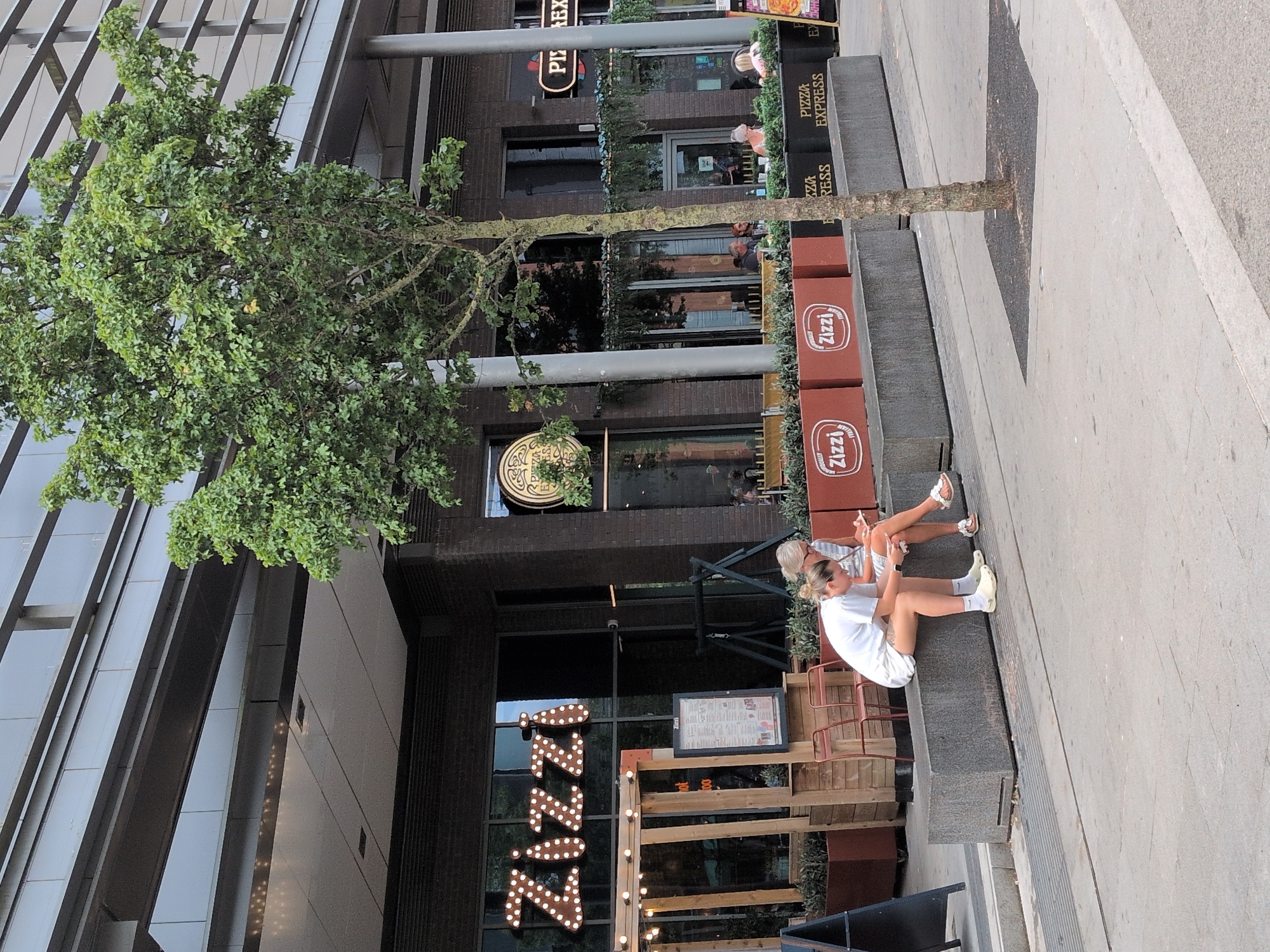 Two young women are sitting on a grey concrete bench outside a Zizzi restaurant and a Pizza Express restaurant. The woman on the left is wearing a white t-shirt and white shorts, and the woman on the right is wearing a white and grey striped dress. Both women are looking at their phones. The Zizzi restaurant has large illuminated letters on its facade. A small tree is planted in a square of dark asphalt in front of the Pizza Express restaurant. Several potted plants and outdoor seating areas are visible in front of both restaurants. The buildings have a modern architectural style with large windows and metal framing. The scene appears to be in a public outdoor area in a city, likely Telford, United Kingdom, given the presence of these well-known restaurant chains.