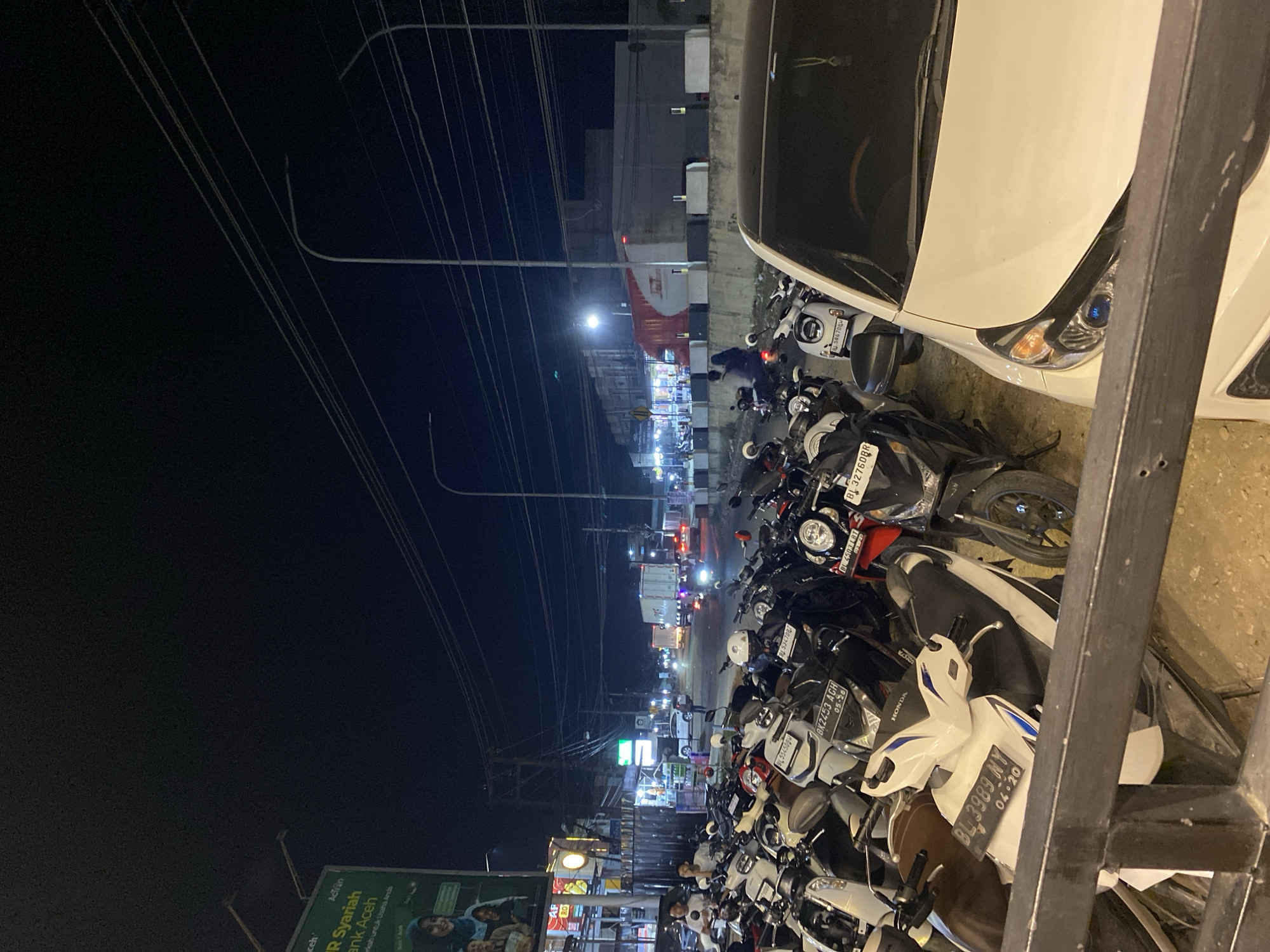 At night in Seuneubok Teungoh, Indonesia, a densely packed row of motorbikes lines a street. A white car is parked to the right, partially obscuring the view. Utility wires crisscross overhead. The scene is illuminated by streetlights and the headlights of distant traffic. A billboard featuring the text "R Syariah Bank Aceh" is visible on the left.