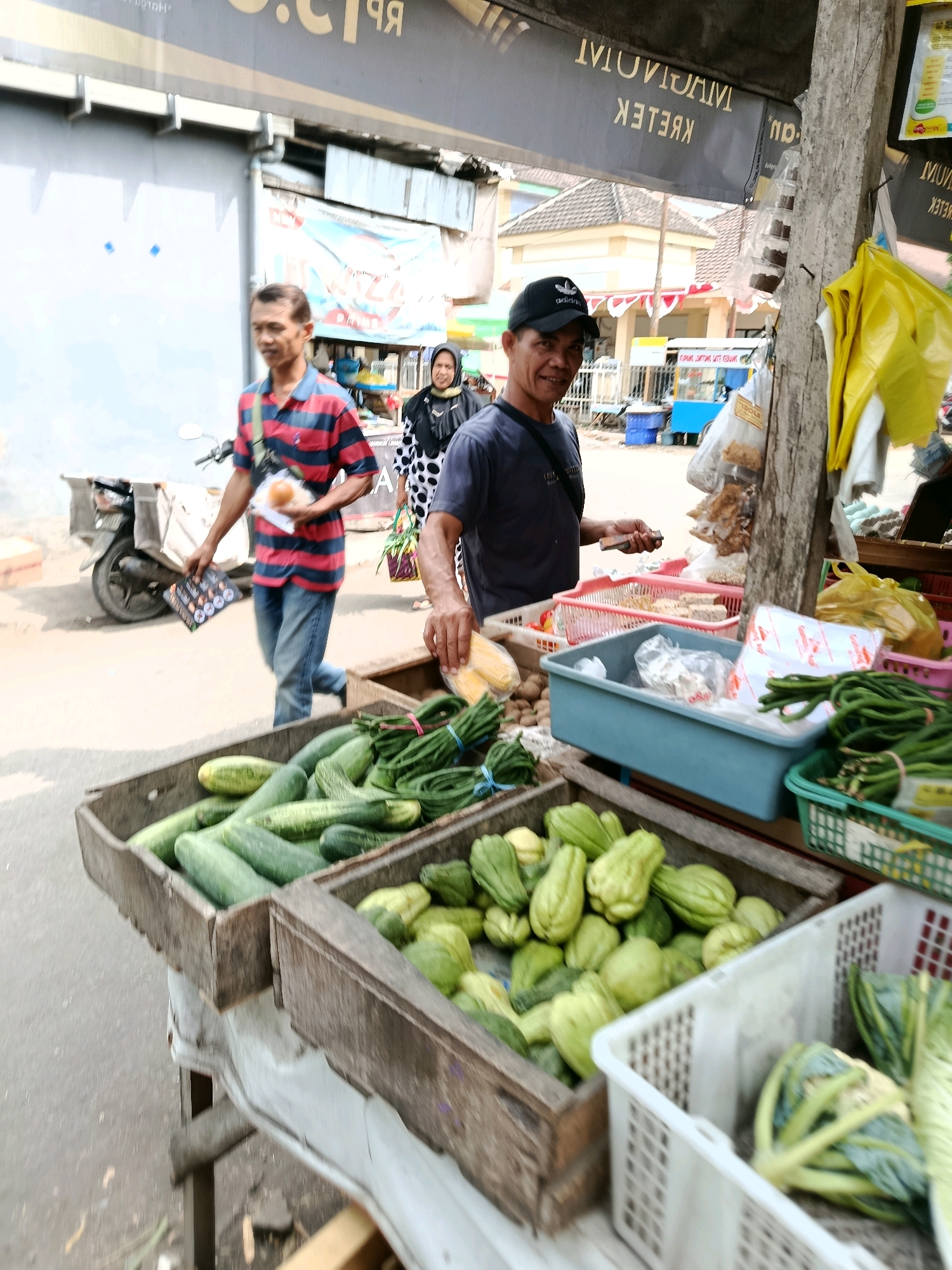 A street market scene in Bululawang, Indonesia, shows a vendor attending to customers. In the foreground, several crates overflow with fresh produce. One wooden crate is filled with cucumbers, while another, also wooden, contains a large pile of chayote. A white plastic crate to the right holds what appears to be cabbage or cauliflower. In the background, a man in a red and blue striped shirt walks away from the camera, holding a bun and a pamphlet. Another man, wearing a black cap and a dark t-shirt, stands behind a stall and smiles at the camera. He is holding a small object, possibly a phone, in his right hand. A woman in a black and white polka dot hijab walks past in the middle ground. Various other market stalls and awnings are visible, with signs indicating the location. The overall atmosphere suggests a busy local market on a bright, possibly warm day.