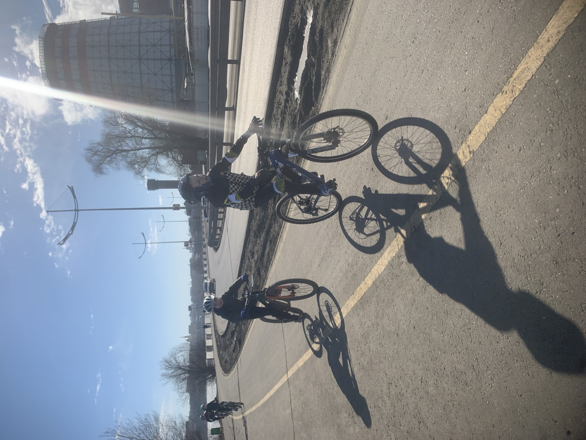 The image shows a group of cyclists riding on a paved path in Izhevsk, Russia. In the background is a tall industrial structure. Two cyclists are prominently visible in the foreground. The cyclist on the right is wearing a checkered pattern shirt, helmet, and cycling gear, and is actively riding. The cyclist on the left is wearing similar gear, including a helmet, and is also riding. Shadows of the cyclists are cast on the pavement, indicating strong sunlight. The path is marked with a yellow line. Further back, there are other cyclists and indistinct urban elements. The environment appears to be a mix of urban infrastructure and open space, possibly along a river or other waterway. The weather appears to be clear and sunny.