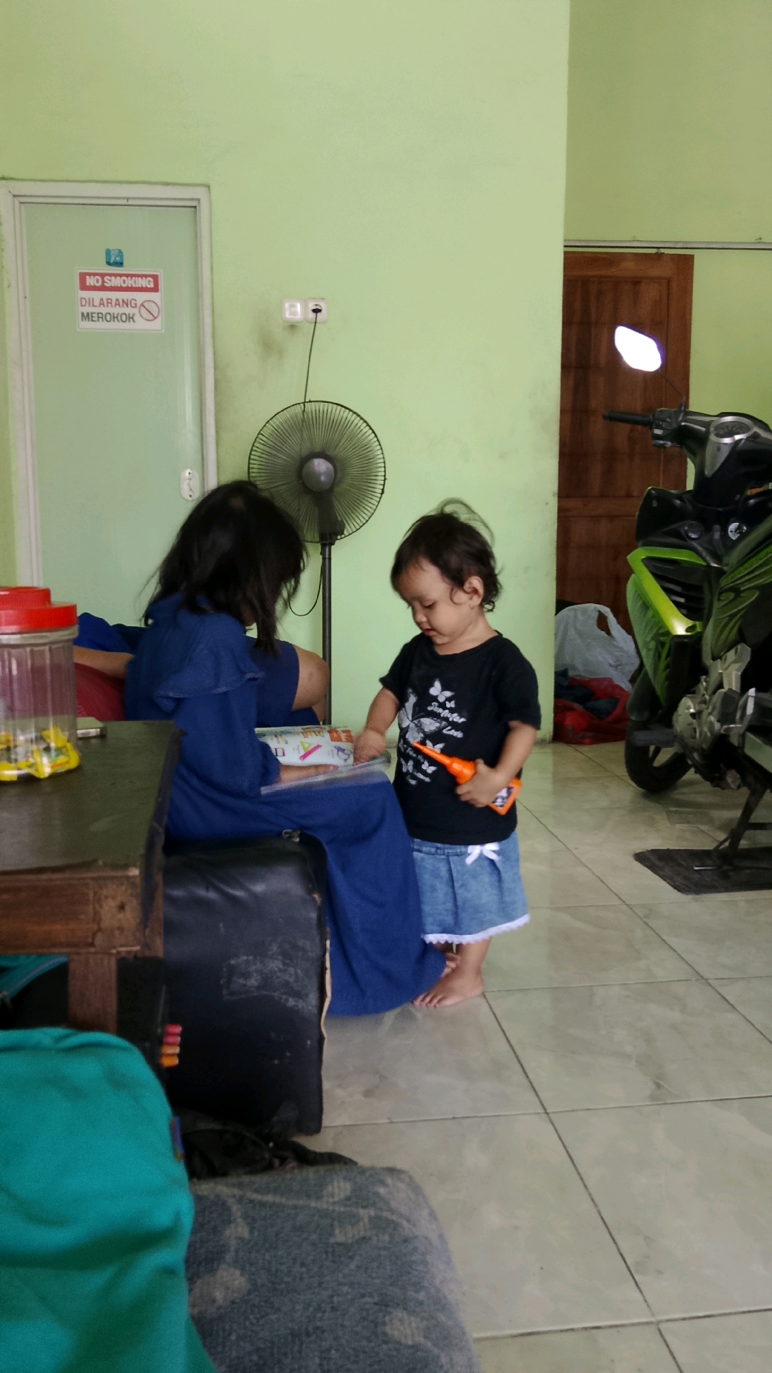 In Sukorame, Indonesia, a young child wearing a black t-shirt and a denim skirt stands on a tiled floor, holding an orange toy. To the left, an older child or adult, dressed in a blue garment, is seated and appears to be reading. A fan is visible behind them. The room has light green walls and features a closed door with a "No Smoking" sign. In the background, a motorcycle is parked, and a wooden door is visible. The scene is indoors, likely in a residential or common area.