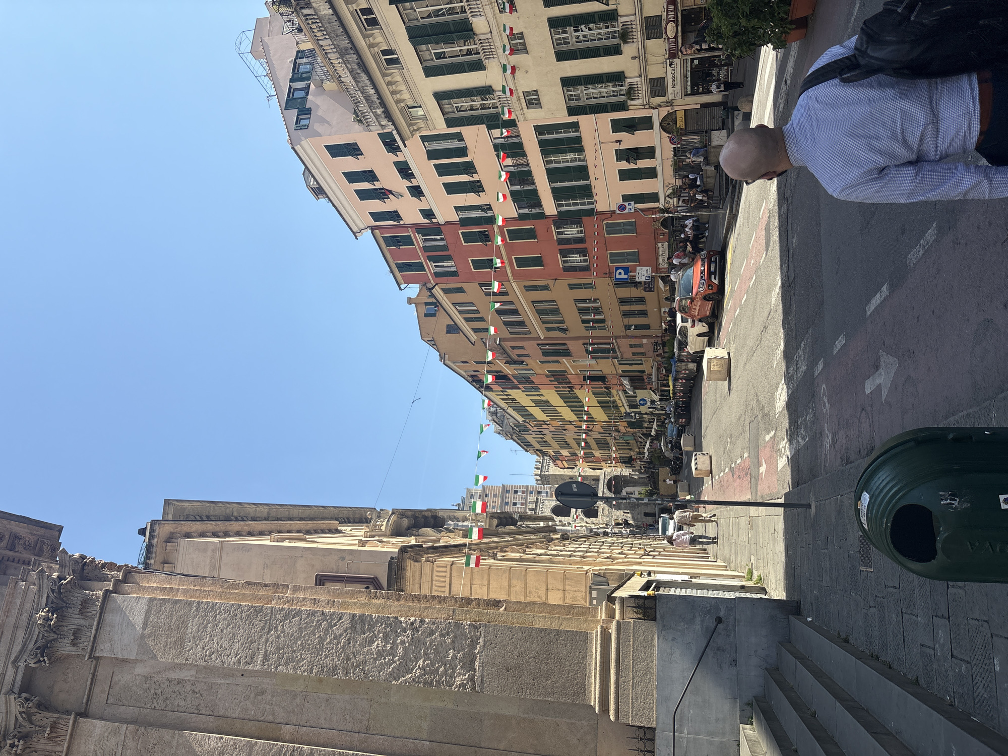 The image captures a street in Genoa, Italy. Buildings line both sides of the street, decorated with Italian flags strung overhead. A bald man wearing a light blue shirt and a backpack is partially visible on the right. In the foreground on the right side of the image, a green trash bin sits on a sidewalk made of gray paving stones next to a set of stairs. Cars are parked along the street. People are walking in the distance. The sky is clear and blue.