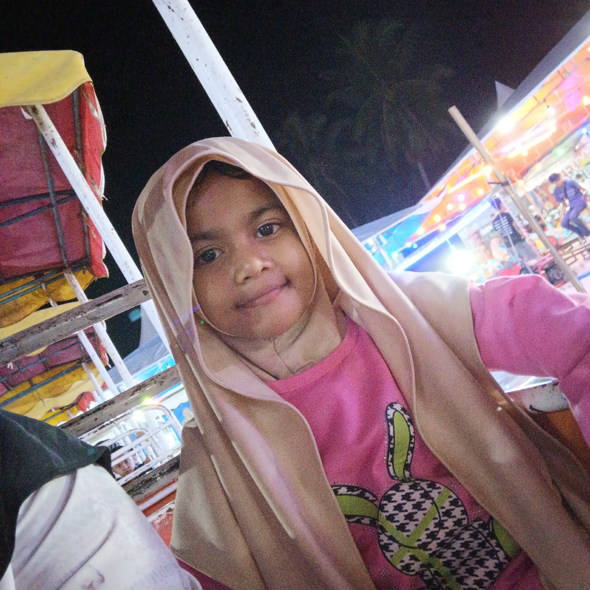A young girl wearing a pink shirt with a cartoon bunny design and a tan hijab poses for a selfie at night. She is seated, and the background suggests she is at an amusement park or fair, with colorful lights and structures visible. To her left, there are red and yellow awnings and white poles, characteristic of carnival rides. In the distance, more rides with bright lights and people can be seen. The image is taken at a slightly upward angle, focusing on the girl's face and upper body.