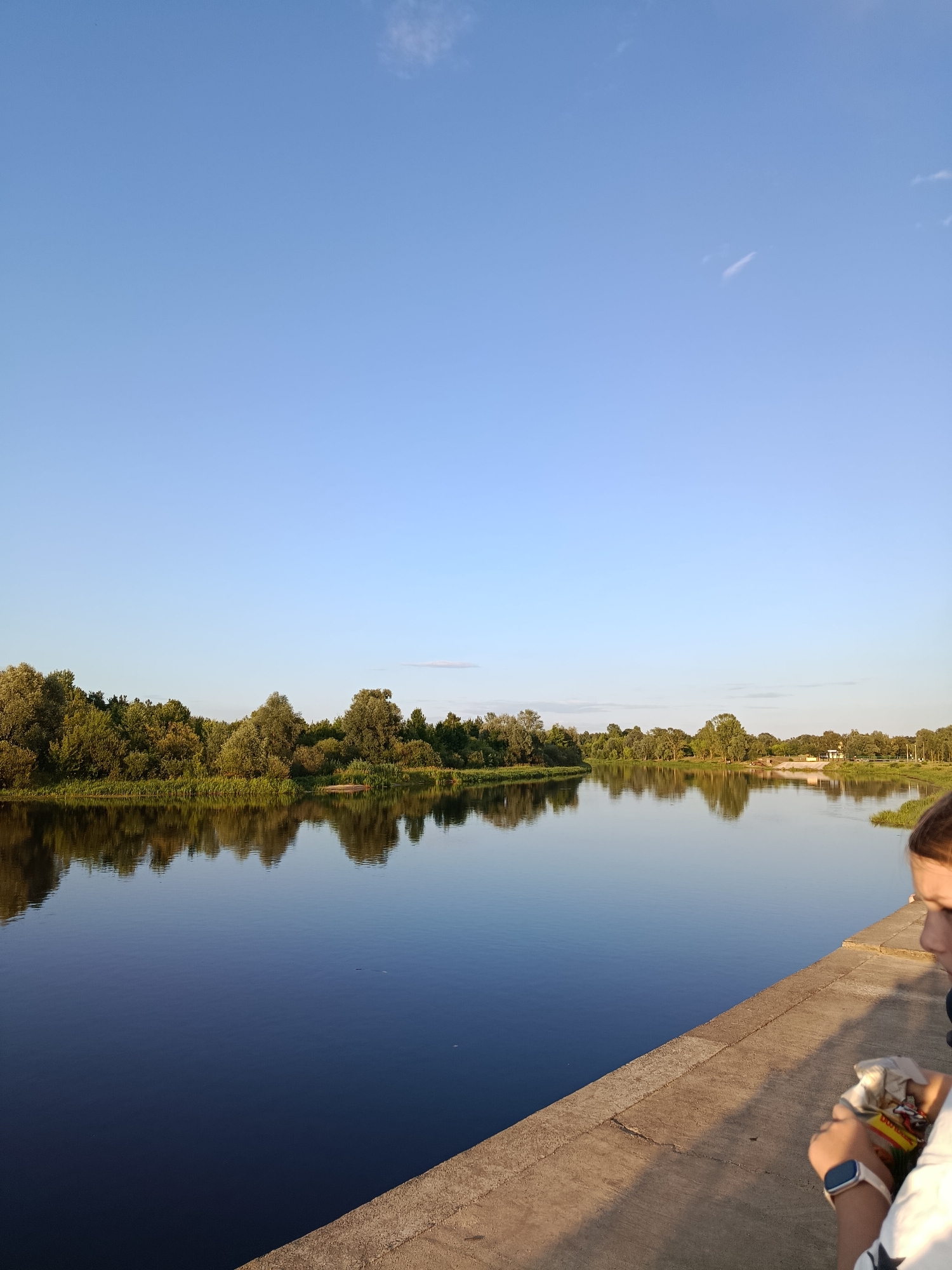 The image depicts a wide, dark blue river with calm waters reflecting the clear blue sky and a lush green treeline on the opposite bank. The treeline is dense with various shades of green foliage. In the foreground, a concrete embankment runs diagonally from the bottom right corner of the frame. A person, partially visible on the embankment, is shown from the shoulders down, looking to the left. They are wearing a white shirt with dark stars and a white smartwatch on their left wrist. The person is holding a white wrapper and appears to be interacting with something within it. The overall scene suggests a peaceful outdoor setting, likely during the late afternoon or early evening due to the soft lighting. The location is identified as being in the city of Svietlahorsk, Belarus.