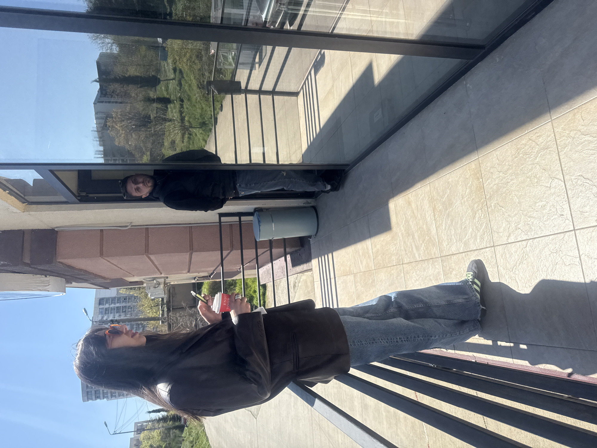 The image shows a woman standing on a balcony in Tbilisi, Georgia, holding a cup and a phone. She is wearing a dark blazer, jeans, sunglasses, and dark sneakers. The balcony has a metal railing and tiled flooring. In the reflection of a nearby window, a man is seen wearing a black jacket and jeans. The reflection also shows a view of buildings and trees. The sky is clear and blue. There is a cylindrical object behind the woman, likely a ventilation shaft or utility fixture.