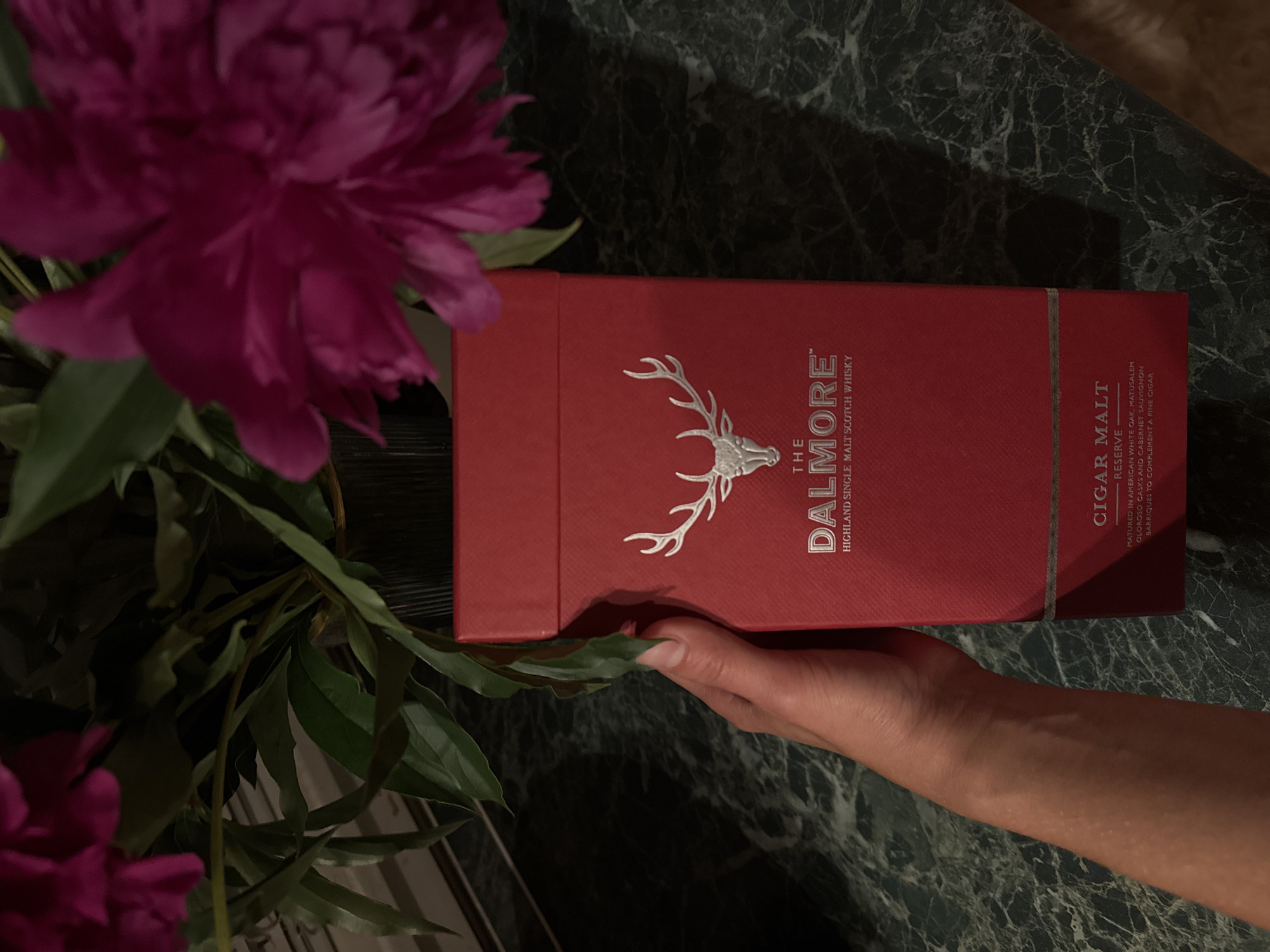 In a dimly lit room with a dark green marble countertop, a hand is holding a red box with a silver stag logo and text that reads "THE DALMORE HIGHLAND SINGLE MALT SCOTCH WHISKY CIGAR MALT RESERVE". The box is positioned next to a bouquet of pink peonies. The scene does not provide any information that would allow for its location to be determined to be in the city of Chaikovsky, Russia.