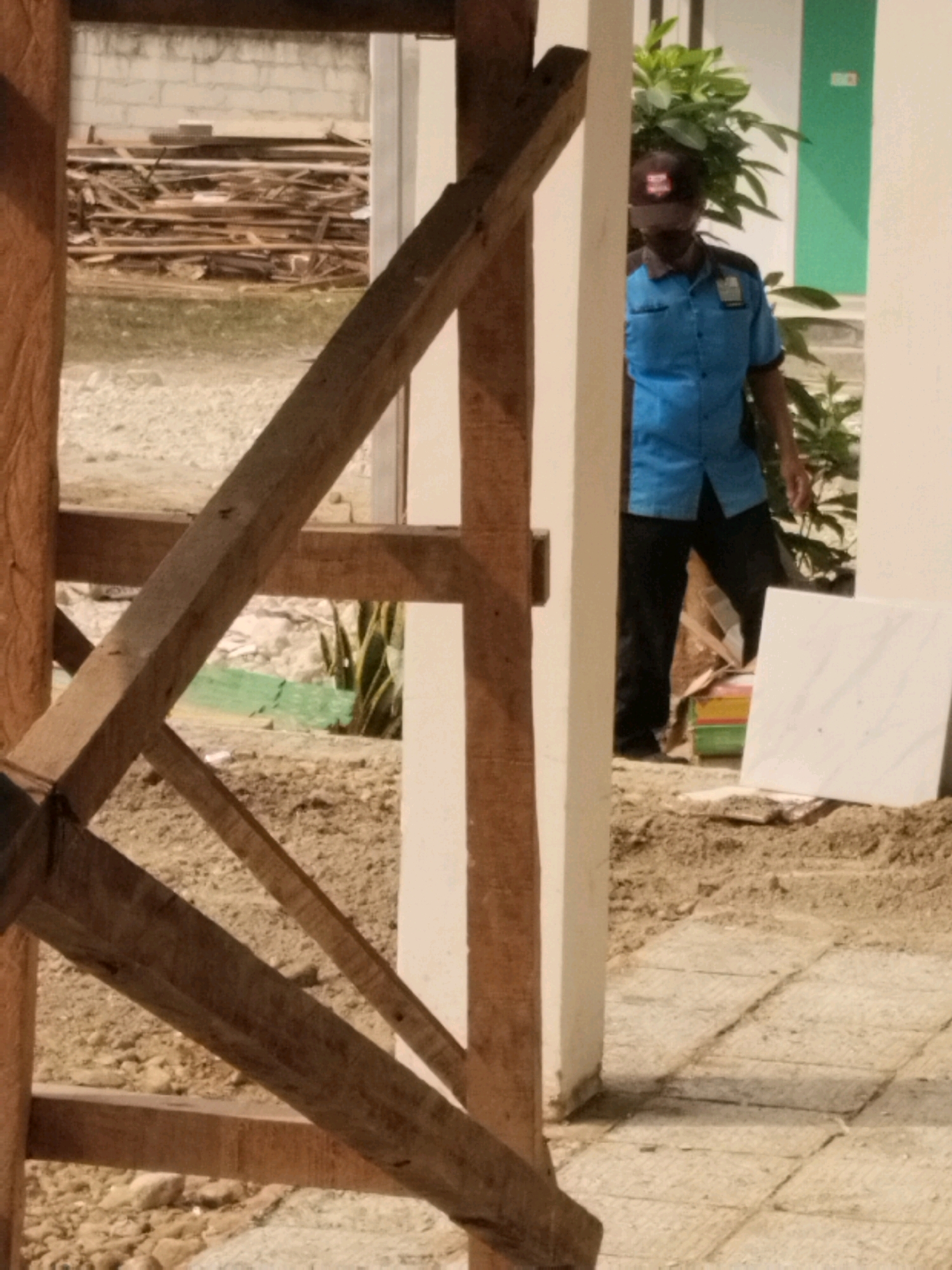 The image depicts a person wearing a blue shirt and black pants, a dark baseball cap, and a face covering, standing near a potted plant. The person appears to be a security guard due to their attire and cap. The scene is outdoors with a construction setting, indicated by piles of wood and construction materials. There is a white tiled surface and loose soil on the ground. The background features a white wall and a green wall, possibly part of a building. The image appears to be taken in Serang, Indonesia, given the context of the provided information.