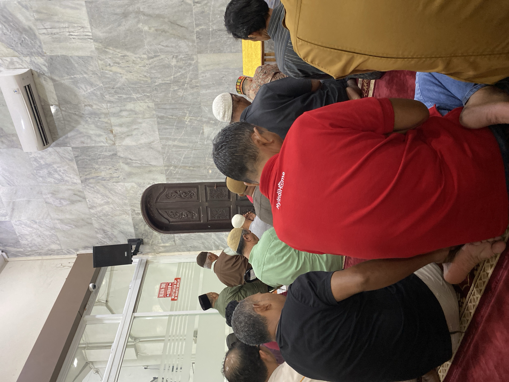 The image depicts a group of men praying inside a mosque. The men are dressed in casual attire, mostly shirts and pants, and some are wearing traditional caps. They are seated on prayer mats, facing a large wooden door. The walls of the mosque are covered in marble-patterned tiles, and an air conditioning unit is visible on the upper right wall. A sign with red lettering is affixed to a glass partition on the left side of the image, which appears to be a rule for the mosque. The overall scene suggests a moment of communal prayer within a mosque in Lhokseumawe, Indonesia.