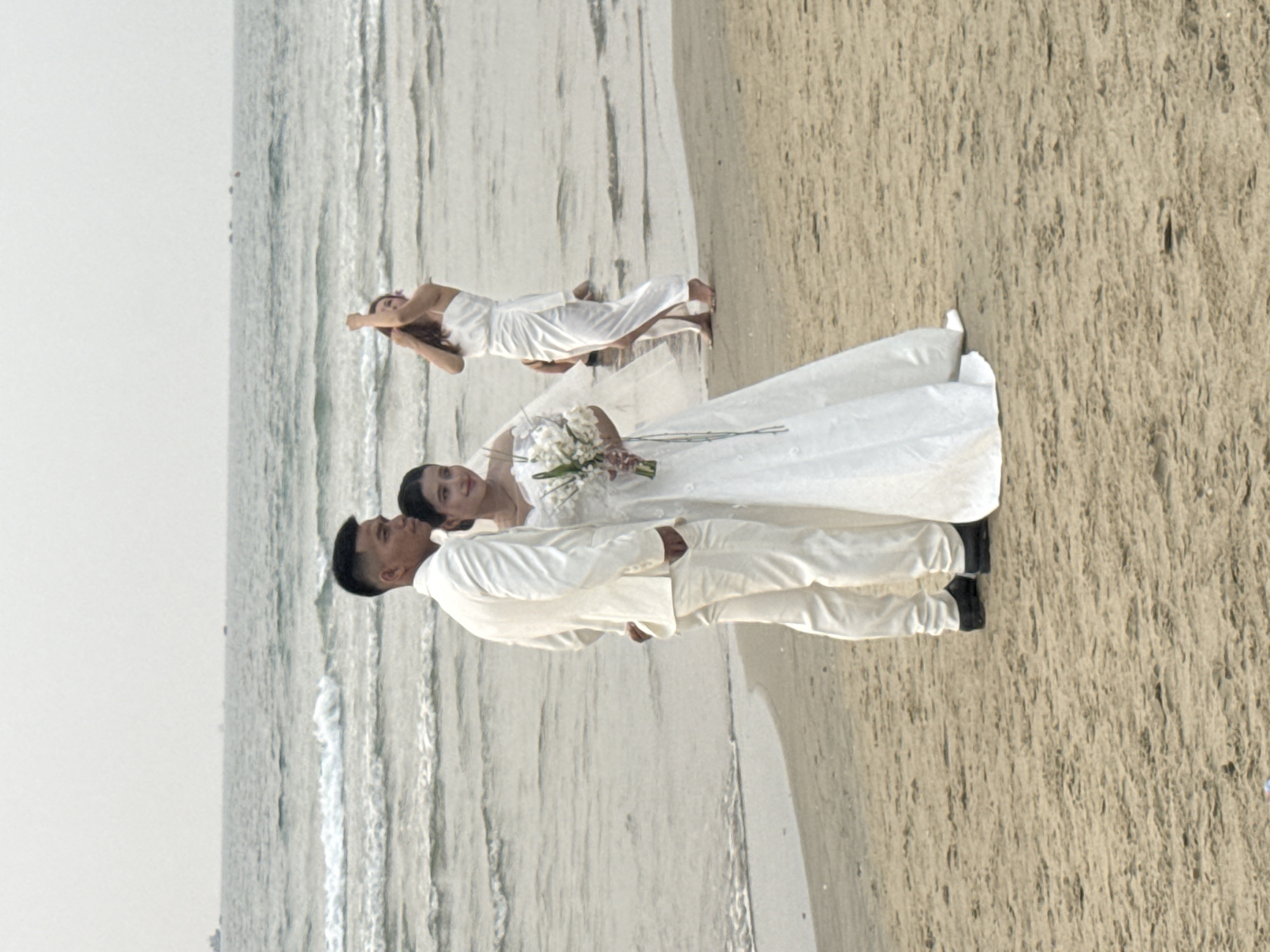 A wedding photograph depicts a bride and groom standing on a sandy beach next to the ocean. A woman is lying on the sand in the background. The scene takes place in Đà Nẵng, Vietnam. The groom wears a white suit and black shoes. The bride wears a long white wedding dress and holds a bouquet of white flowers. The sky is light gray.