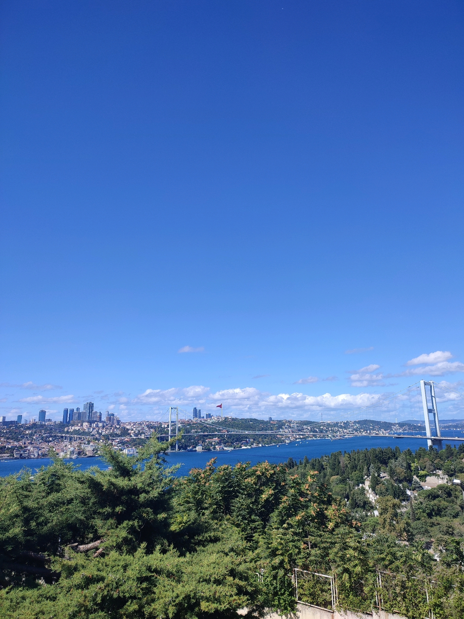 The image captures a panoramic view of the Bosphorus Strait in Istanbul, Turkey, as seen from Üsküdar. The foreground is dominated by lush green trees, with some branches extending into the frame. Beyond the trees, the Bosphorus waterway stretches across the middle ground, reflecting the bright blue sky dotted with scattered white clouds. A suspension bridge, identified as the Bosphorus Bridge (also known as the 15 July Martyrs Bridge), spans the strait, with its distinctive white towers and cables clearly visible. On the far side of the strait, the cityscape of Istanbul unfolds, featuring a cluster of modern skyscrapers and a denser arrangement of residential buildings and structures. A Turkish flag, a vibrant red with a white crescent and star, is visible atop a hill on the cityscape side, near the bridge. The overall scene is serene, bathed in sunlight, and indicative of a clear, pleasant day. No people or specific actions are discernible.