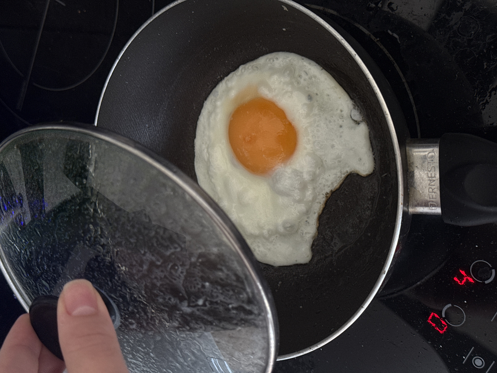 A top-down shot shows a fried egg in a small, black non-stick pan with an "ERNESTO" branded handle, sitting on a black stovetop. The egg has a vibrant orange yolk surrounded by cooked white. A hand holds a clear glass lid slightly ajar, positioned to the left of the pan. The stovetop beneath the pan appears slightly wet. Digital elements on the stovetop indicate numbers "0" and "4" illuminated in red. The image does not provide information about the location in Vozdovac Urban Municipality, Serbia.