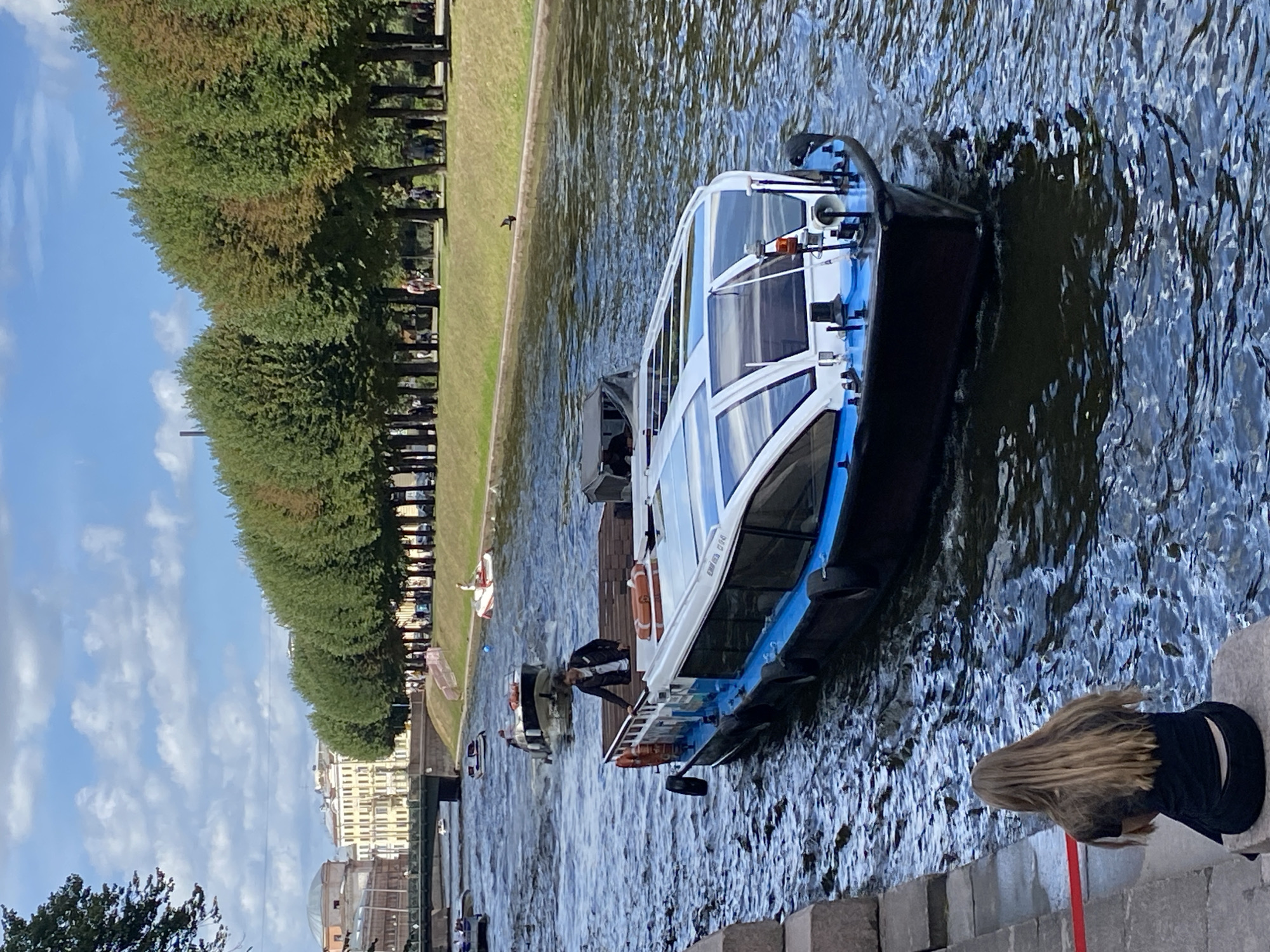 The image depicts a canal in Saint Petersburg, Russia, with a tour boat in the foreground. The boat is white and blue with a glass enclosure and is moving through the water. A person is standing on the deck of the boat. On the left bank of the canal, a person with long blonde hair is sitting on the stone embankment. The right bank of the canal is lined with a row of trees, behind which are buildings and a bridge in the distance. The sky is partly cloudy. Several other smaller boats are visible in the canal further down.