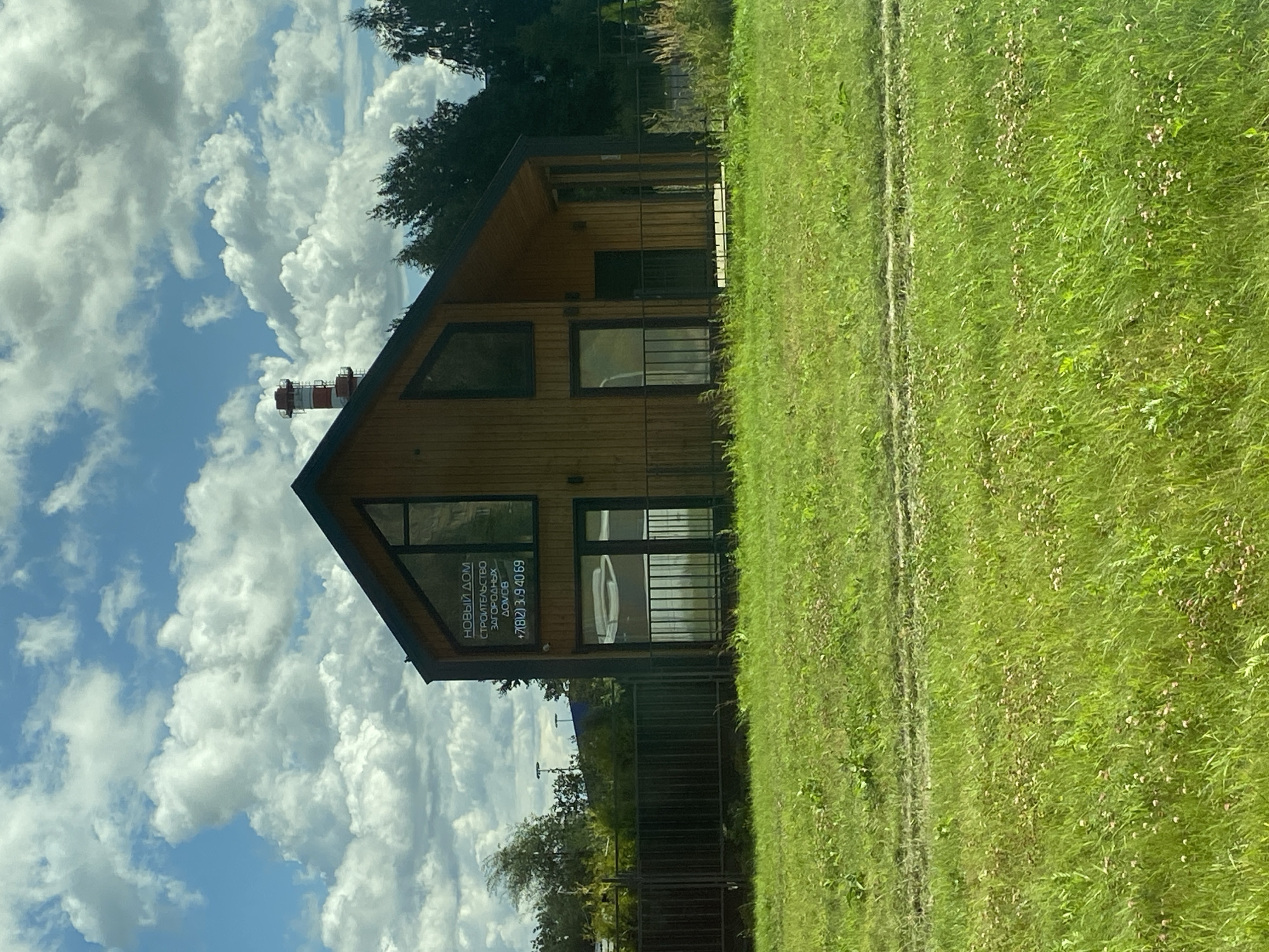 A modern wooden house with a dark roof and large windows stands in a grassy field under a cloudy blue sky. A red and white striped industrial chimney is visible behind the house to the left. The house has a sign on the front with Russian text and a phone number. A dark metal fence encloses the property in front of the house. The grass in the foreground is green and somewhat overgrown, with scattered small pink flowers. The overall scene suggests a suburban or rural setting on a partly cloudy day.