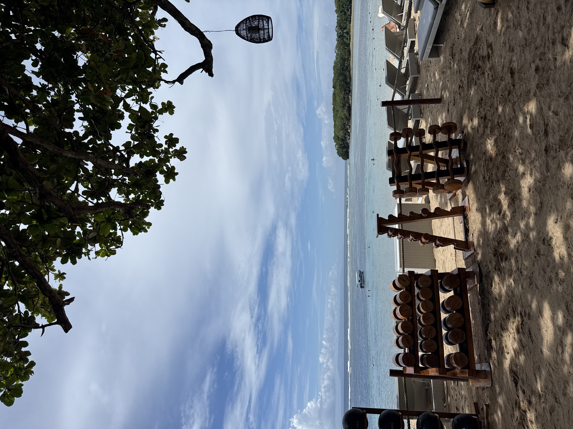 The image captures a beachside scene, likely in Kuta Selatan, Indonesia, given the tropical foliage and coastal environment.
On the right side of the frame, there are several racks of wooden dumbbells, typical of an outdoor fitness setup. Their arrangement suggests a workout area set up directly on the beach. Shadows indicate strong sunlight, likely during the daytime. The ground is sandy, confirming the beach location.
In the background, there is an ocean view with what appears to be a distant shoreline or island. Several lounge chairs are set up on the beach, suggesting a resort or recreational area. A single boat can be seen in the distance.
On the left, there is a leafy tree branch overhanging the scene, contributing to the tropical ambiance. A decorative, woven-style light fixture hangs from the tree branch. The sky above is blue with scattered clouds.