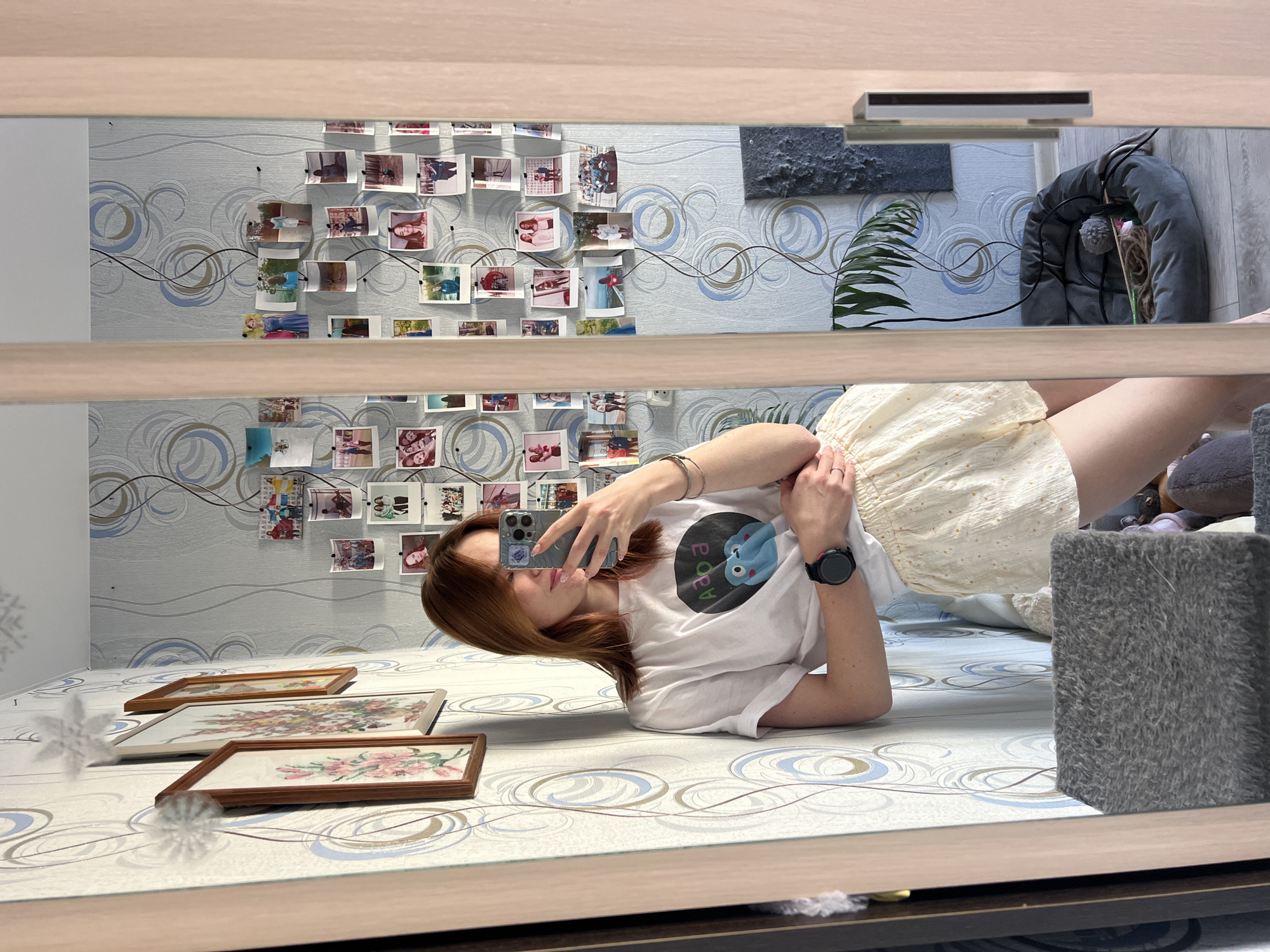 A woman is taking a selfie in a mirror. She is lying on a surface with a patterned design, presumably a bed. Three framed pictures are arranged in front of her. The wall behind her is decorated with patterned wallpaper and a collage of photographs attached by pins or tacks, resembling a decorative display. A plant and a pet bed are partially visible in the background. The location is Chaikovsky, Russia, based on prior information.