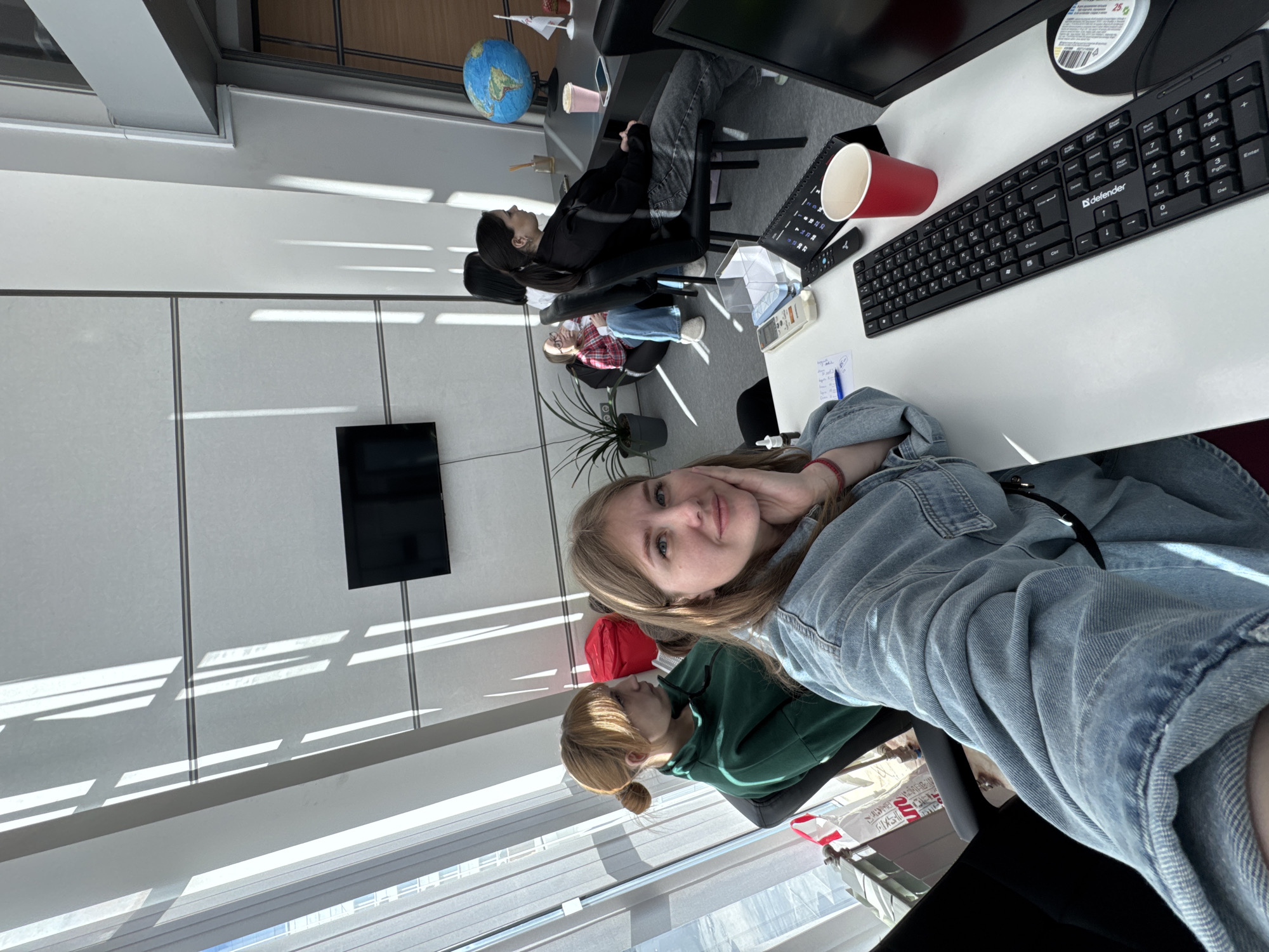 A woman is taking a selfie inside an office space. The location is Perm, Russia. The woman has blonde hair and is wearing a denim jacket. Behind her, another person is wearing a green sweater and a red hat. Several other people are seated at desks working, and office equipment like keyboards, monitors, and a globe are visible. Sunlight is coming through the windows. There's a wall-mounted television and an indoor plant.