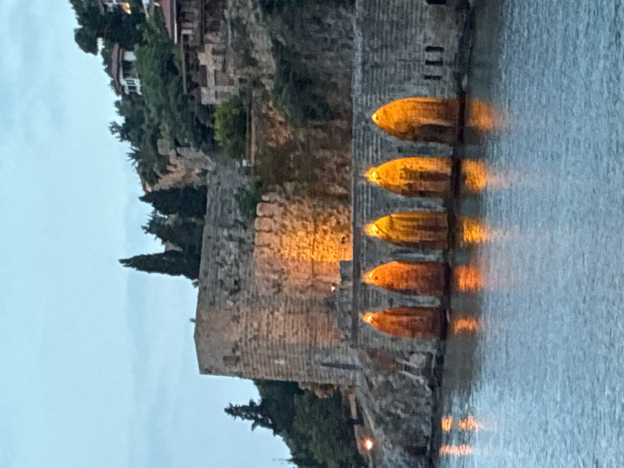 The image captures a tranquil scene at dusk or night along a coastline, likely in Alanya, Turkey. On the left, a robust, ancient stone wall, possibly part of a fortress or castle, rises from the water's edge and extends up a hillside. This hillside is densely covered with dark green trees and foliage. Further up and partially obscured by greenery, faint outlines of buildings or houses are visible.
A series of five bright, golden-orange lights are vertically arranged along a dark, sturdy structure that extends into the water. These lights cast a warm glow on the adjacent stone wall. The calm body of water to the right prominently displays distinct, elongated, oval-shaped reflections of each light source. The sky is a dark, clear blue-grey, indicating either late dusk or nighttime, with artificial lights illuminating the scene. The overall impression is one of historical ambiance and serenity, with no human activity or visible text.