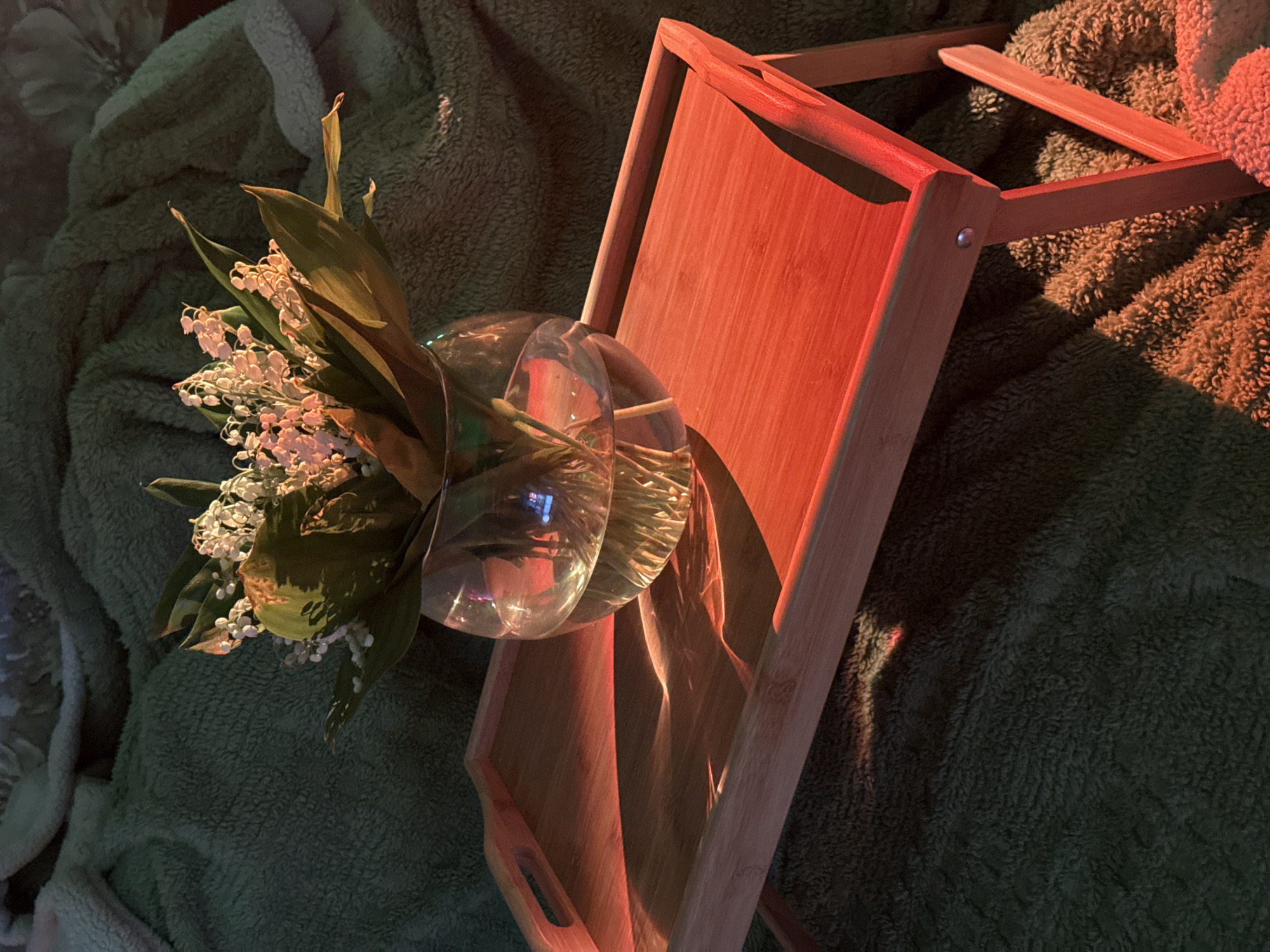 The image shows a glass vase filled with lily of the valley flowers resting on a wooden serving tray. The tray is placed on a soft, plush blanket. The lighting is predominantly warm, with red and orange tones casting shadows across the scene. The exact location within Unknown, Unknown is not discernible from the visual information provided.