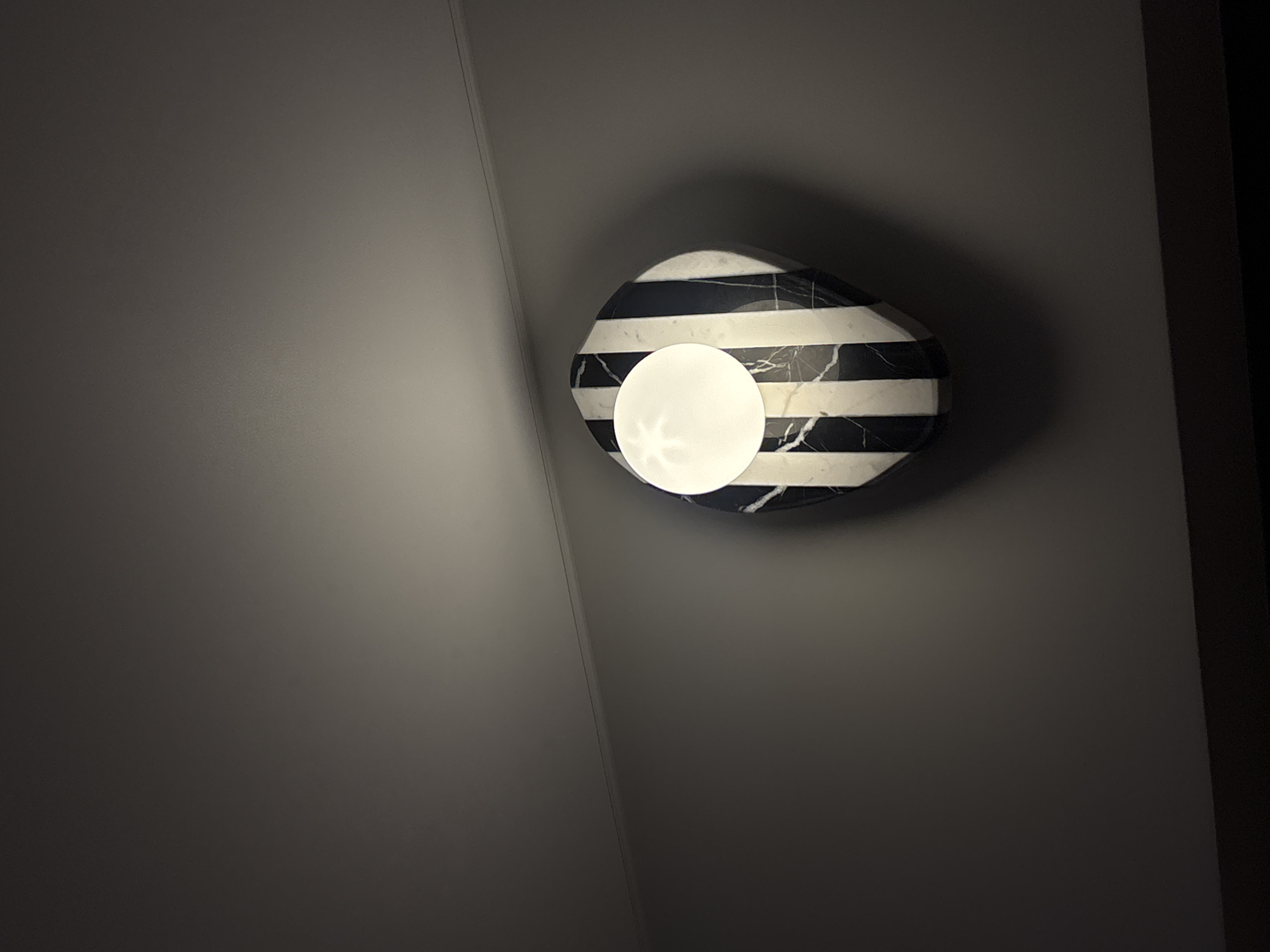 A wall-mounted light fixture is affixed to a gray wall. The fixture is an oval shape with horizontal black and white stripes. A white, circular light emanates from the center. The wall appears to be indoors and is located in Krasnoyarsk, Russia.