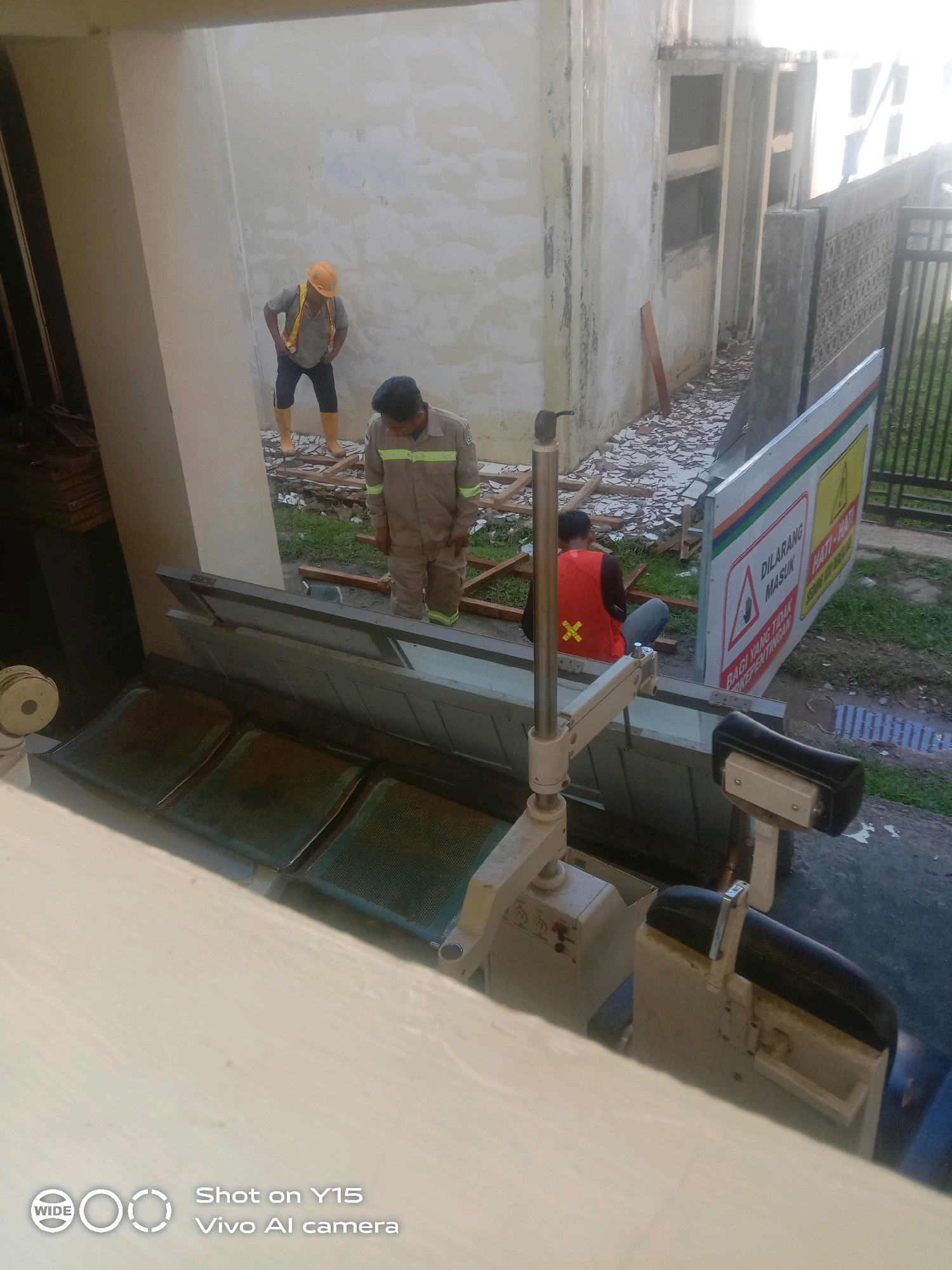 In Manyang Cut, Indonesia, a scene of ongoing construction or renovation is depicted. Two male workers are present. One, wearing a yellow hard hat and reflective vest over a grey shirt and dark pants, stands near a light-colored wall, appearing to be observing or waiting. The second worker, dressed in a tan jumpsuit with reflective stripes and a black shirt, is crouched down, possibly working on debris on the ground. In the foreground, a metal structure with green seating is visible, possibly an outdoor seating area or a disassembled bench. Behind the seated worker, a large sign with red and yellow warning symbols and text in Indonesian, including "DILARANG MASUK" (No Entry), is prominently displayed. The environment appears to be outdoors, with scattered debris and some greenery visible on the ground. The overall impression is a work-in-progress site with a focus on safety warnings.