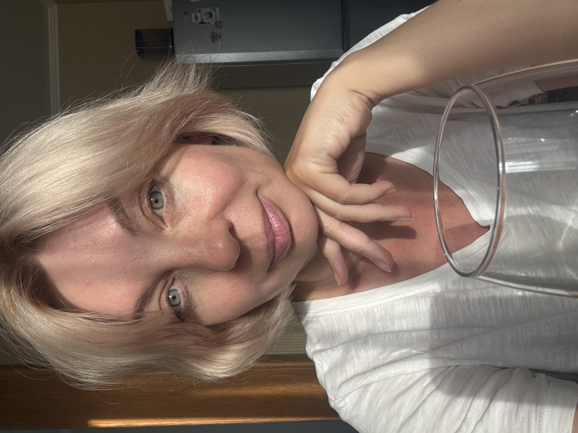 In this close-up, front-facing portrait, a woman with blonde, chin-length hair and light blue eyes is positioned in the foreground. She is wearing a white t-shirt and resting her chin on her hand, with her fingers extended. A clear glass is placed in front of her. The lighting suggests it is daytime, with sunlight casting shadows from the left side of the frame. The background is blurred, showing hints of a domestic interior, possibly a kitchen, with a refrigerator visible on the right. There is no discernible location in Sochi, Russia.