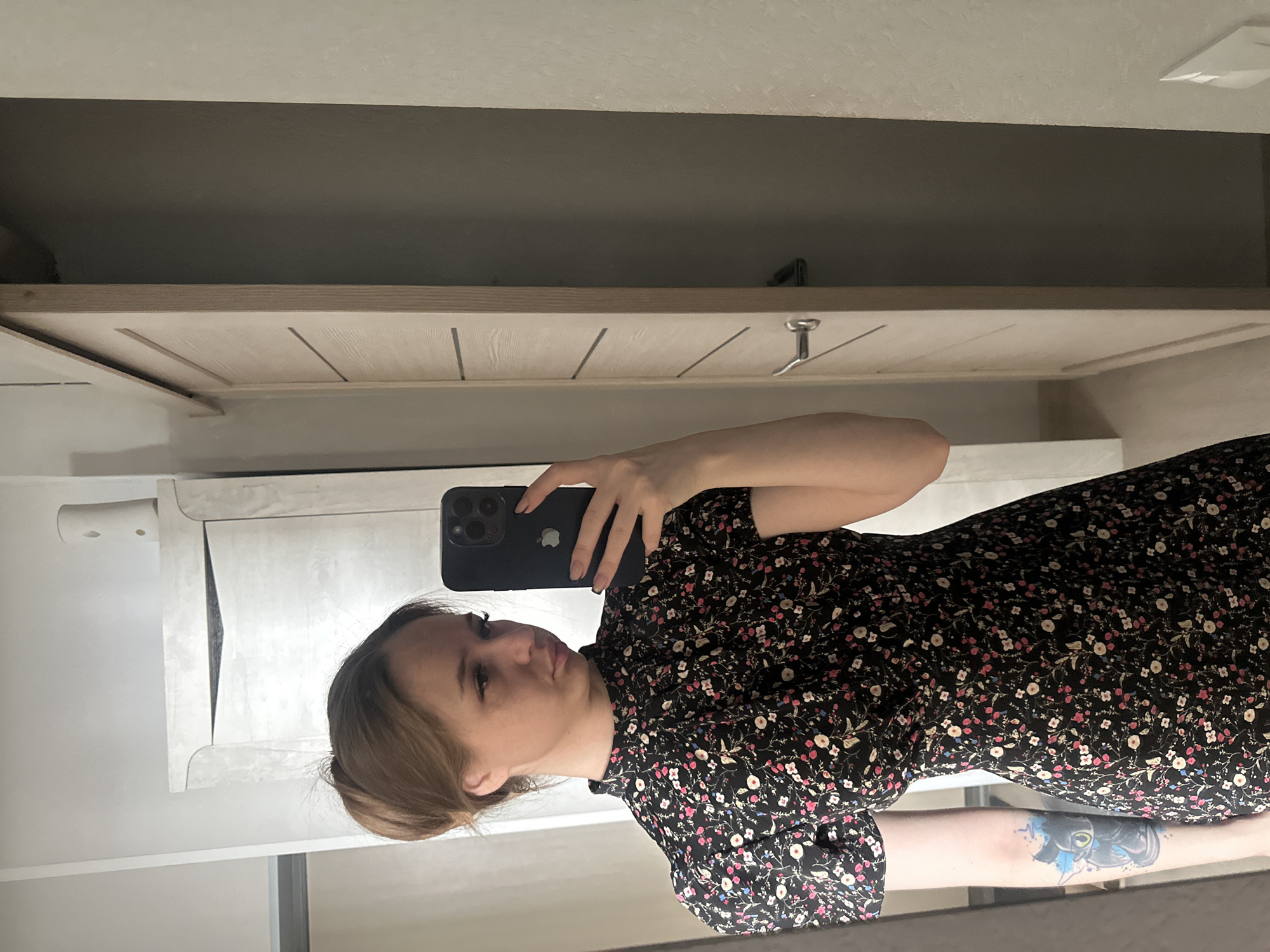 A young woman with light brown hair pulled into a bun stands in what appears to be an indoor setting, reflected in a mirror. She holds a dark-colored smartphone, possibly an iPhone due to the visible Apple logo on the back, up to her face, capturing her reflection. Her left arm is visible holding the phone, while her right arm is angled downwards, revealing a colorful tattoo on her forearm depicting a black cat-like creature with blue accents. She is wearing a dark-colored dress adorned with a pattern of small pink, white, and blue flowers, featuring short puffy sleeves and a high neckline. Her lips are slightly pursed, and her gaze is directed towards the phone.
The background visible in the mirror shows a light-colored wall. To the left of the woman's head, an air freshener canister is visible on top of a light-colored, ornate piece of furniture or cabinetry. To her right, a light-toned wooden sliding door with horizontal grooved lines is partially open, revealing a dark interior space. A metallic door handle is visible on the sliding door. The floor appears to be a light-colored surface, possibly wood or laminate. The lighting suggests an indoor environment, likely a well-lit room.