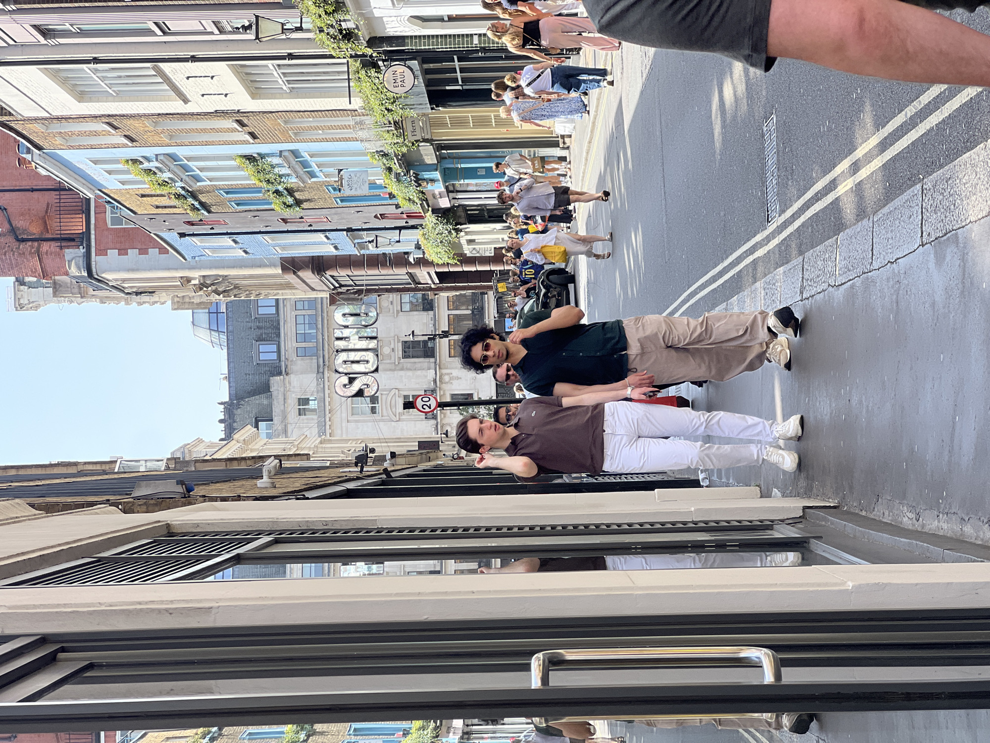 The image shows a street scene in the Soho area of London, United Kingdom. Two men are standing on the sidewalk near a storefront with a large window displaying reflections. One man is wearing a brown polo shirt and white pants, while the other man wears a dark green shirt and khaki pants. Behind them, the street is filled with pedestrians walking in both directions, and on the other side of the street, storefronts line the sidewalk. Signs indicating "SOHO" are visible. A speed limit sign showing "20" is seen. The architecture features a mix of building styles, with varying heights and facades. The weather appears sunny.