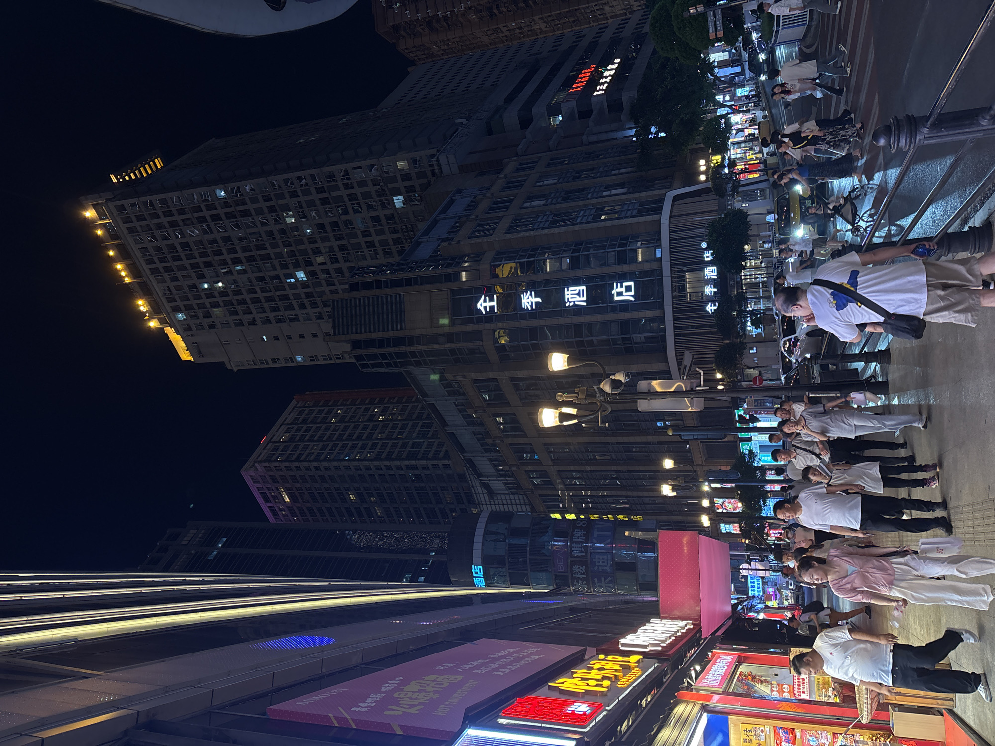 Here is a description of the image:
The image presents an evening cityscape within the Yuzhong District of Chongqing, China, oriented with an unusual tilted perspective. Tall buildings flank a commercial street bustling with pedestrian traffic. Numerous lights illuminate the street, highlighting storefronts adorned with signage in both Chinese characters and English lettering. One building displays the Chinese characters "全季酒店" (Quanji Jiudian), which translates to "JI Hotel" in English.
Several groups of pedestrians are present, appearing to move along the sidewalk. The image provides a candid snapshot of urban life with a focus on the architectural elements, signage, and movement within the scene.