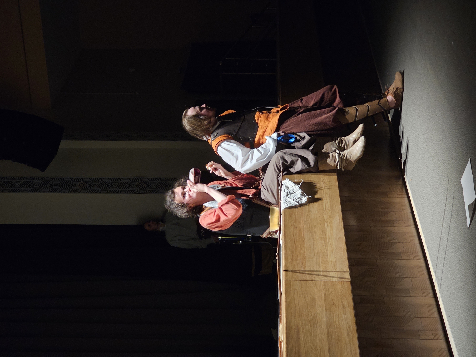 The image depicts a theatrical scene on a wooden stage in what is assumed to be the city of Tosno, Russia. Two individuals are prominently featured, dressed in period costumes. On the left, a person with curly hair and glasses, wearing an orange tunic and a dark vest, is seated and appears to be drinking from a cup. To their right, another individual, with medium-length brown hair, is standing and looking to the right, clad in a white shirt, an orange vest with dark trim, and brown trousers with leather armor on the lower legs. A third person, partially obscured in the background on the left, stands in a light-colored tunic against a dark curtain. The stage is made of light-colored wood, with a dark, patterned panel along the lower front edge. The background is dark, with hints of stage lighting and equipment. The ground in front of the stage is a textured grey surface.