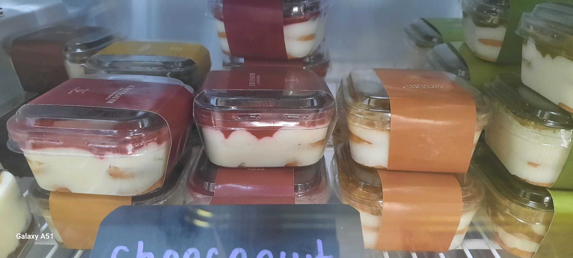 The image shows a close-up view of several plastic containers of desserts stacked inside a refrigerated display case. The desserts appear to be layered, with a white cream-like substance, possibly cake or biscuit layers, and fruit or sauce toppings. Some containers have labels with text, including "RED VELVET" and "CARAMEL". The display case is brightly lit, and the refrigeration unit is visible in the background. The overall impression is one of a retail setting, likely a bakery or dessert shop. The text at the bottom left corner of the image "Galaxy A51" indicates the phone used to take the picture. There is no information about the city of Beru, Indonesia, or any people or actions within the image.