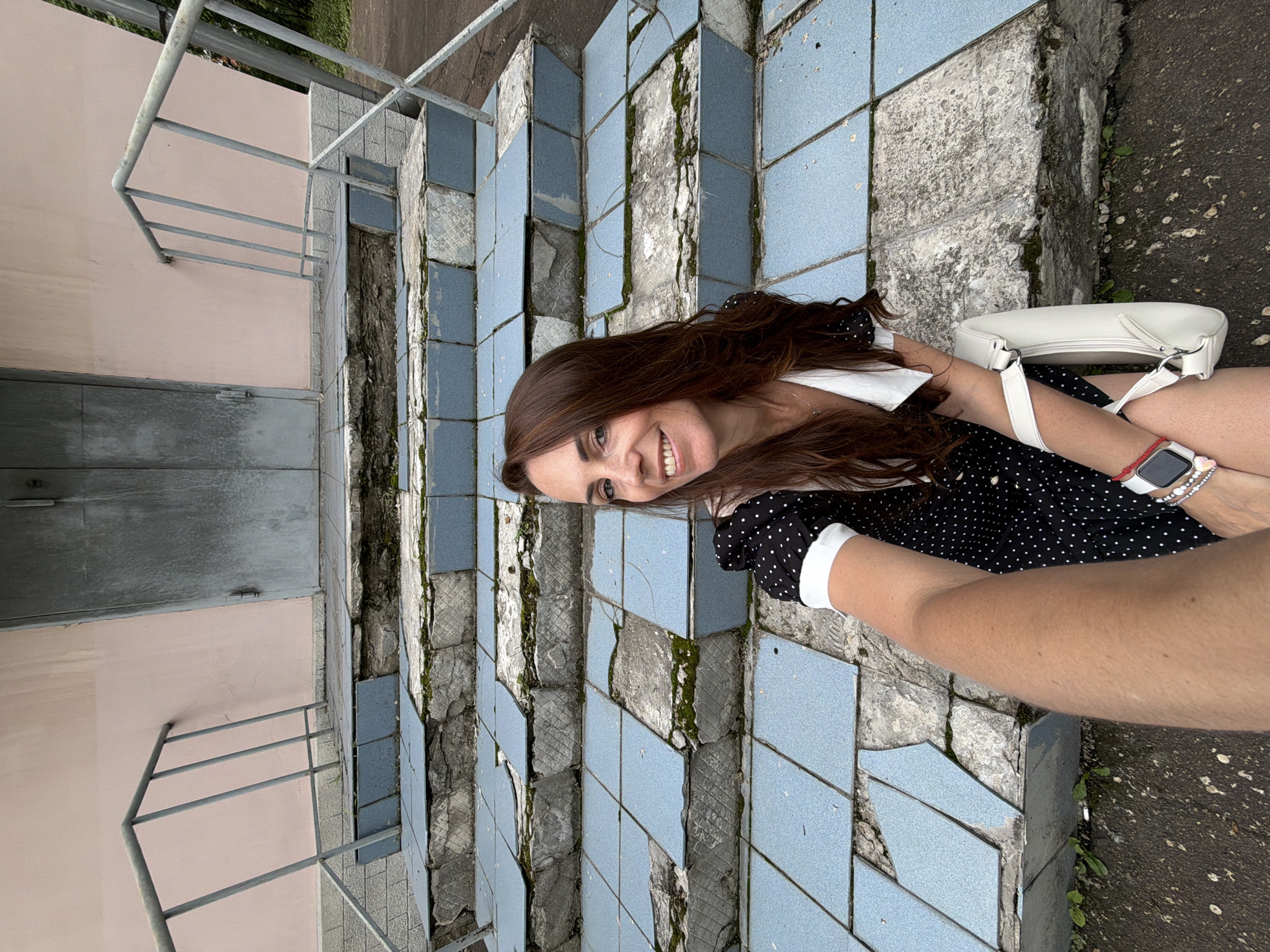 A woman with long, dark brown hair, wearing a black dress with white polka dots and a white collar, is seated on a damaged outdoor staircase. She is holding a white handbag and has a smartwatch on her left wrist. The staircase is covered in blue tiles, many of which are broken and missing, exposing the concrete underneath and patches of moss. The building behind her has a pale pink wall and a large, weathered grey metal door. Metal railings run along the sides of the stairs. The image appears to be a selfie. The location is indicated to be in the city of Birobidzhan, Russia.