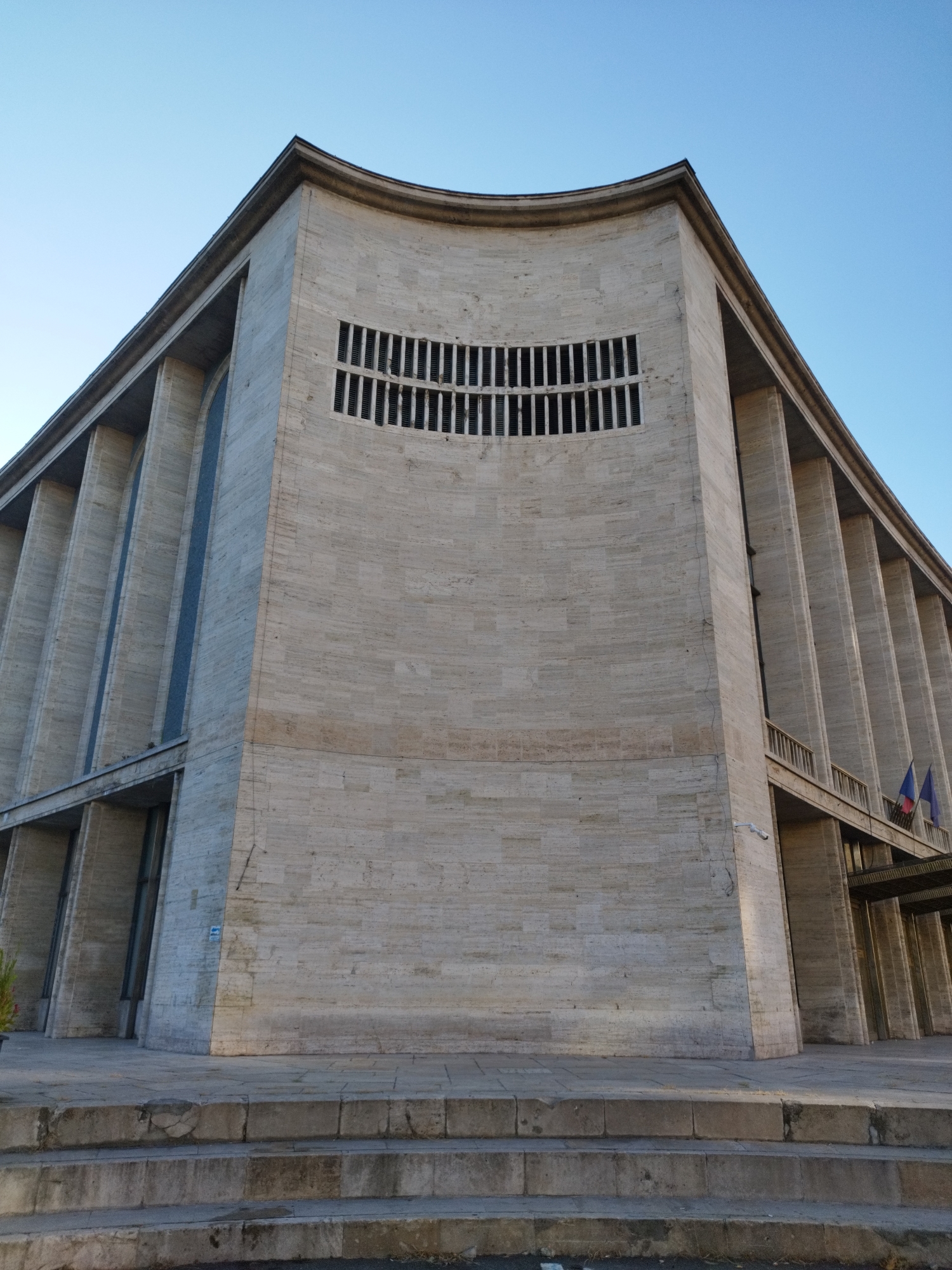 A low-angle, full shot captures a section of the Palace of the Parliament in Bucharest, Romania, under a clear, pale blue sky. The building's facade is constructed of light-colored stone blocks, exhibiting a visible grain. A prominent curved architectural feature dominates the upper portion of the visible facade. Below this curve, a rectangular window with vertical slatted blinds is centered. The building's structure is characterized by a series of vertical, recessed elements separated by pilasters, creating a ribbed effect along the sides.
On the right side of the facade, a balcony with a metal railing is visible, supported by the pilasters. Two flags, one with red, yellow, and blue horizontal stripes and another solid blue, are mounted on poles to the right of the balcony. A security camera is mounted on the wall below the balcony. A horizontal band of architectural detail, appearing to be another balcony or ledge, runs across the lower part of the building's visible section.
In the foreground, a set of stone steps leads up to the building. The steps are worn and appear to be made of large, roughly cut stone blocks. The ground around the steps is paved with stone tiles. The lighting suggests it is either early morning or late afternoon, casting subtle shadows. No people are visible in the frame. The overall impression is one of monumental architecture and clear, open space.