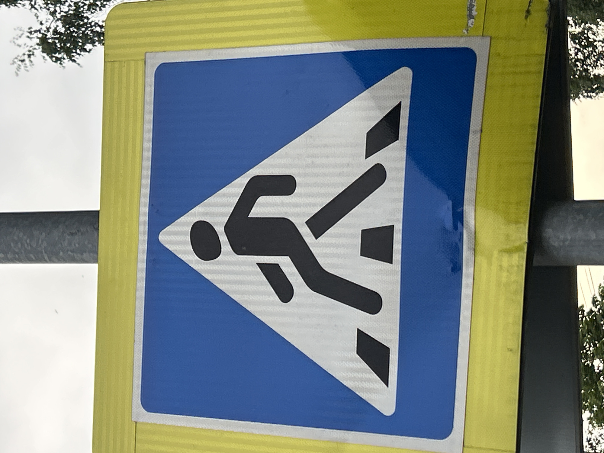 An outdoor shot shows a close-up of a pedestrian crossing sign. The sign is square and yellow with a blue, square inner panel that contains a white triangle with a black silhouette of a person walking. The person is depicted mid-stride on a stylized crosswalk with black rectangular shapes representing the stripes. The sign is mounted on a metal pole. The background consists of a cloudy sky and some blurred trees.
