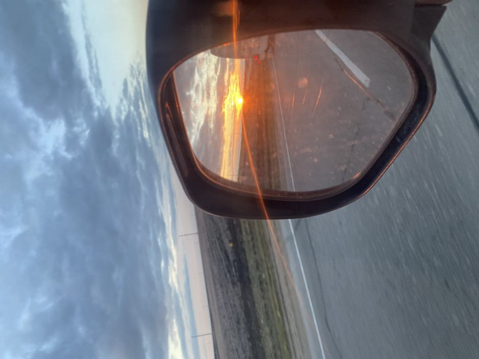 The image presents a view from a vehicle traveling on a multi-lane asphalt road near Prostorny, Russia, at sunset. The left portion of the frame shows the road's shoulder delineated by a solid white line, adjacent to a flat expanse of dark, tilled soil extending to the horizon. Several dark, slender utility poles are visible in the distant background across the field. Above the landscape, the sky is partially covered with grey, broken cumulus clouds, with lighter hues near the horizon.
The right portion of the frame is dominated by a black-cased, exterior side-view mirror. Inside the mirror's reflection, a vibrant orange and yellow sun is depicted low on the horizon directly behind the viewer's vehicle, indicating sunset. Intense orange lens flares emanate from the reflected sun, creating diagonal light streaks that traverse the mirror's surface and extend slightly beyond its frame. The reflection within the mirror also reveals the road surface behind, marked by a white dashed lane line, and a partial view of a trailing vehicle, identifiable by a red taillight on its right side. The mirror's glass surface shows evidence of dust and smudges. No people are visible.