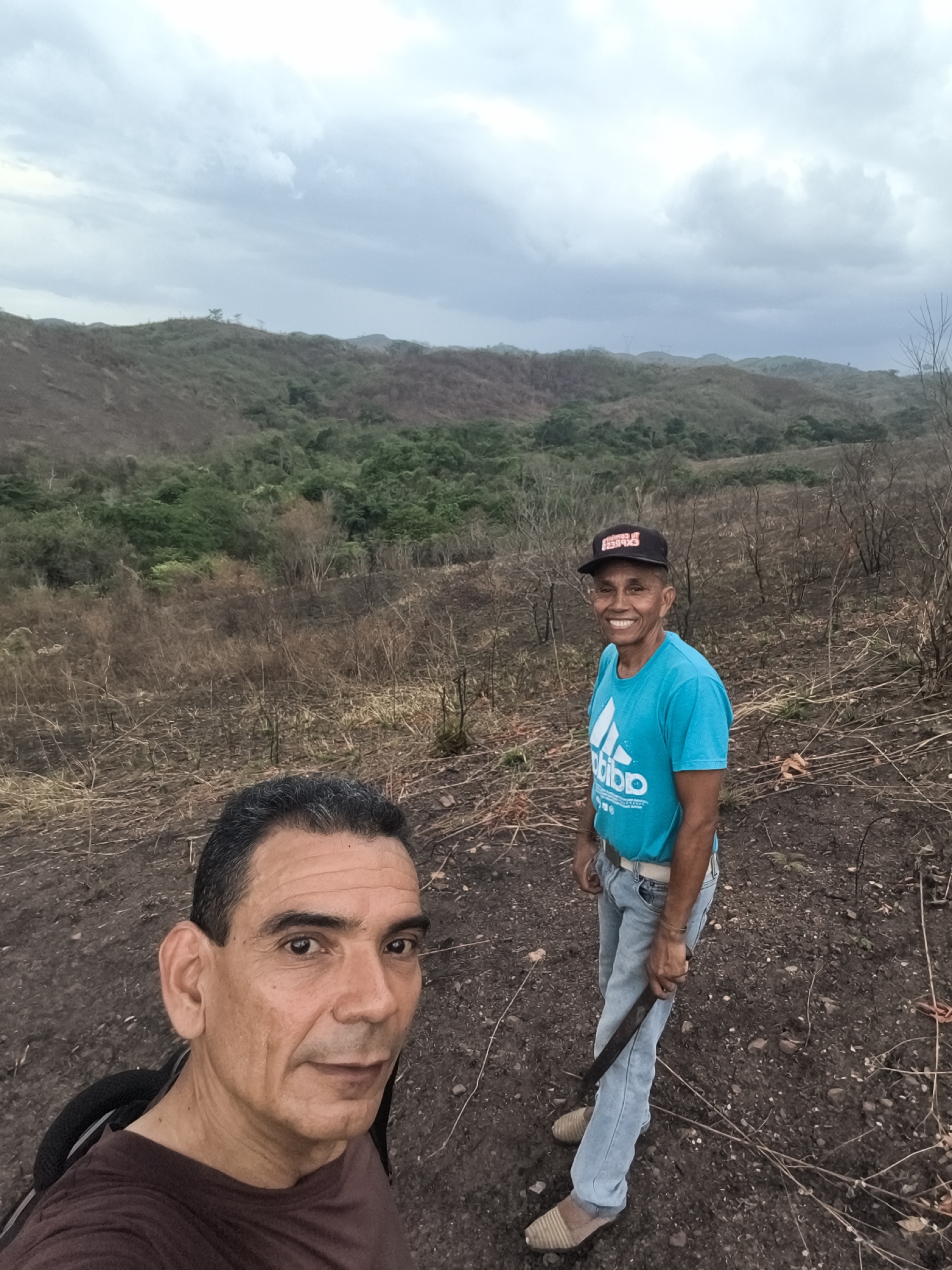 Two middle-aged men are standing in a landscape of dry, brown vegetation and dark soil in Tinaquillo, Venezuela. The man in the foreground, wearing a brown shirt, is taking a selfie. The second man, slightly behind and to the right, wears a blue T-shirt, light blue jeans, and a black cap. He is smiling and holding a machete. In the background, rolling hills covered with sparse vegetation are visible under an overcast sky.