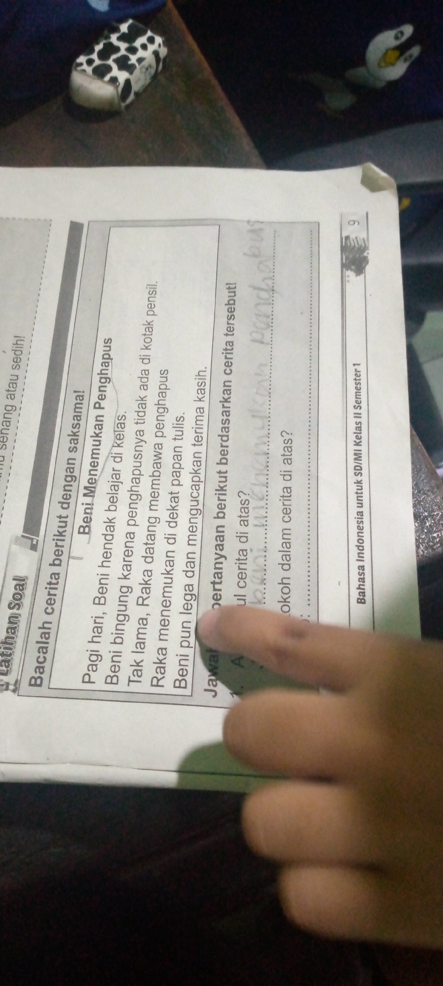 The image is a close-up, angled shot of a page from a textbook or worksheet, likely in Indonesia given the text. The text is primarily in Indonesian and appears to be a reading comprehension exercise. A story about a character named Beni and his friends Raka and Maman is presented, followed by questions. There is also some handwritten text, possibly answers, on the page. The bottom right corner of the page indicates "Bahasa Indonesia untuk SD/MI Kelas II Semester 1," which translates to "Indonesian Language for Elementary School/Madrasah Ibtidaiyah Grade II Semester 1." The background is dark, suggesting the photo was taken in low light conditions, and a person's hand is visible in the foreground, partially obscuring the text.