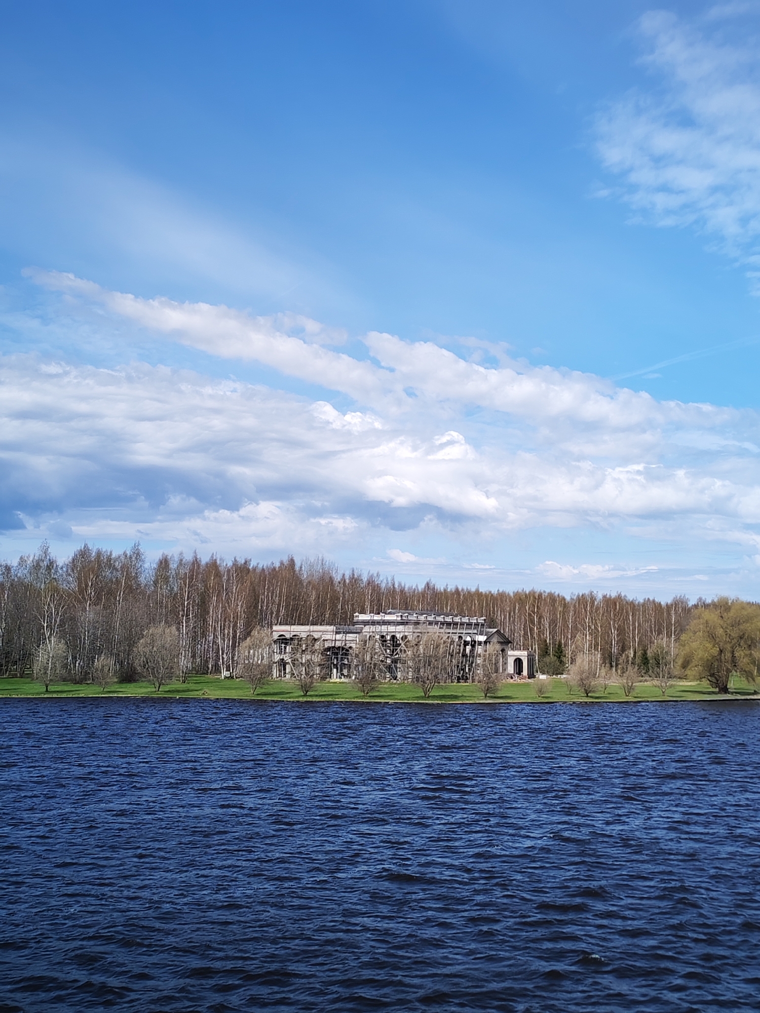 The image shows a body of water in the foreground, likely a lake or a wide river, with gentle waves visible. Behind the water, there is a green strip of land with a building under construction. The building appears to be a large structure with scaffolding visible. It has arched entrances and a partially constructed roof. Surrounding the building is a dense forest with trees that are mostly bare, suggesting it is either early spring or late autumn. The sky occupies the upper portion of the image, with a mix of clear blue and scattered white clouds. There are no people or animals clearly visible in the image.
