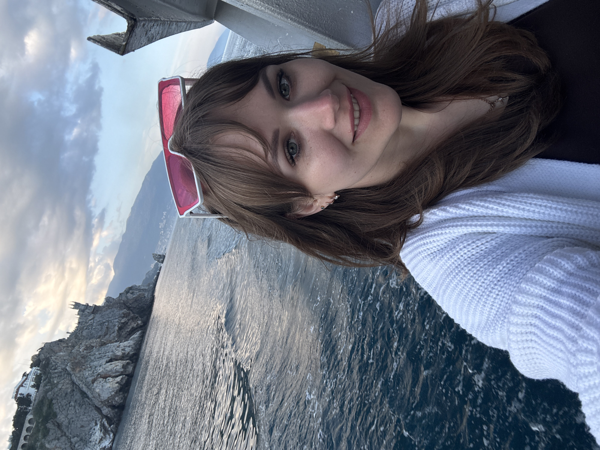 A selfie taken by a fair-skinned woman with brown hair and green eyes, who is smiling slightly at the camera. She is wearing a white cardigan and has pink-framed sunglasses perched on her head. A necklace with a small charm is visible around her neck. The background shows the sea and a rocky coastline with a building atop, suggesting a location in the Гаспринский поселковый совет, Ukraine. The lighting indicates it is daytime, likely early morning or late afternoon.