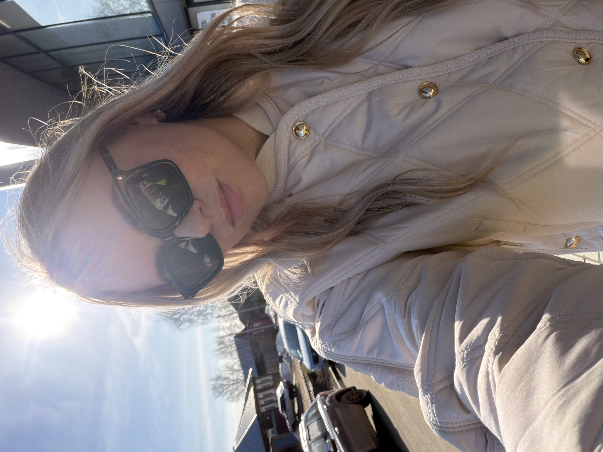 A woman with blonde hair and sunglasses is taking a selfie outdoors on a sunny day. She is wearing a light-colored quilted jacket. Behind her is a street with parked cars and buildings, suggesting a residential or commercial area. The angle of the shot is tilted, capturing the sky and sunlight, which is causing lens flare. It is not possible to verify the specific location of Votkinsk, Russia, from the image provided.