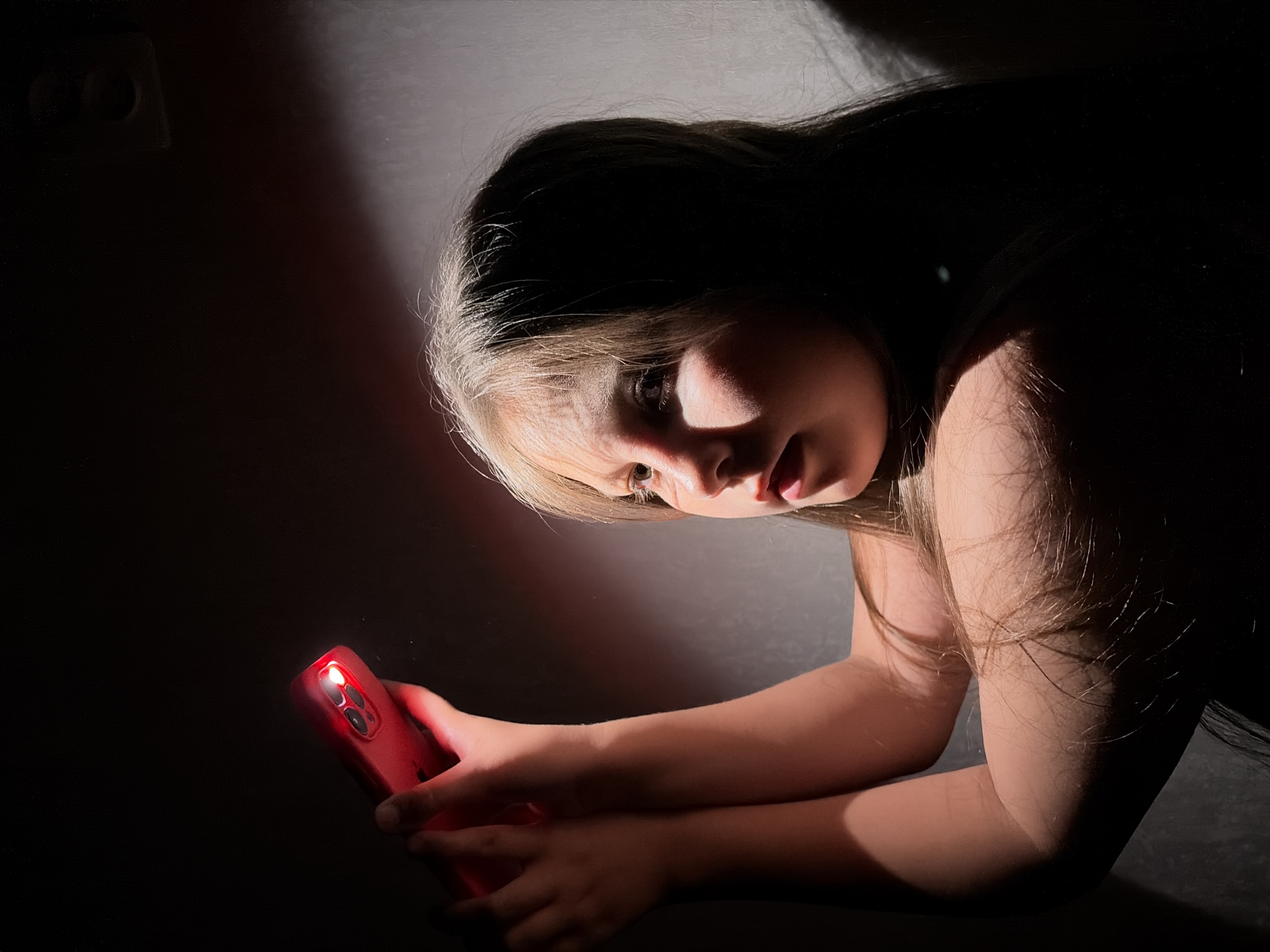 A girl is positioned in a dimly lit room. The only source of illumination is the red light emanating from her smartphone, which she holds in her hands. The phone's glow casts a stark contrast on her face, highlighting her features. Her hair is a mix of dark and light shades. She appears to be focused on something on her phone screen. In the background, a light switch or electrical outlet is visible on the wall. The environment is dark and appears to be indoors. The city is unknown.