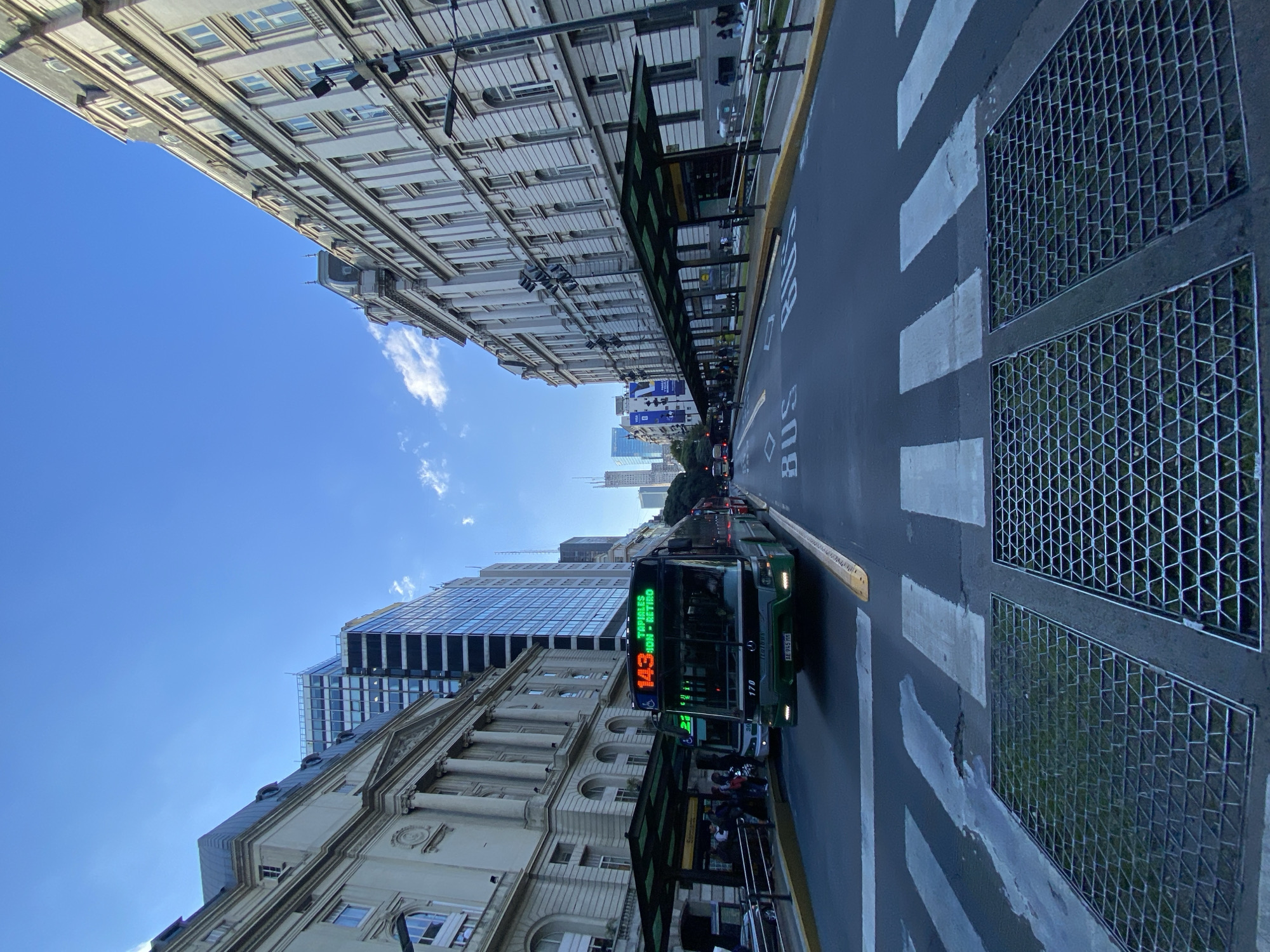 The image captures a street view in Buenos Aires, Argentina. The perspective is from a low angle, looking up between buildings. The sky is blue with a few clouds. On the right, there is a bus lane with a green bus numbered 143, heading away from the viewer. There's a bus stop with people waiting. Pedestrian crossings are marked on the road. Metal grates are visible on the right side of the road. On either side of the road, there are buildings of different heights and architectural styles, including classical and modern designs.